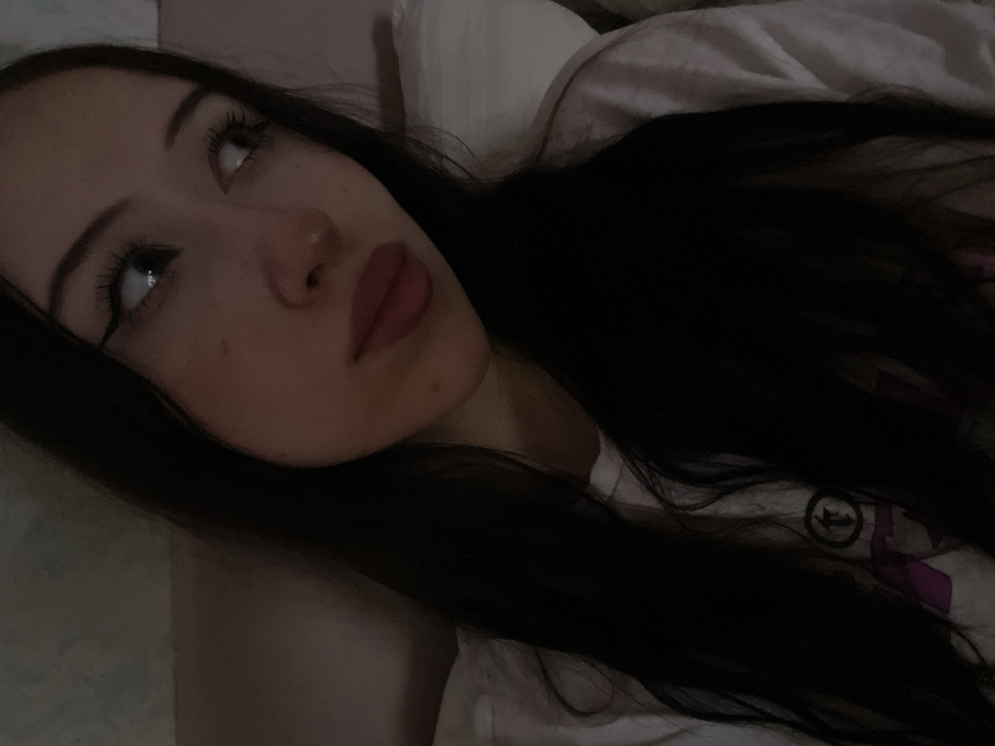 A young woman with long, dark hair and fair skin is lying down, looking off to the right. She is wearing a white t-shirt with a graphic design on it. Her eyes are open, and she has winged eyeliner on. Her lips are full and painted a reddish-pink color. The background appears to be a bed with light-colored bedding. The lighting is dim, suggesting it might be nighttime or a dimly lit room. There are no visible elements in the image that would indicate a specific location within Votkinsk, Russia.