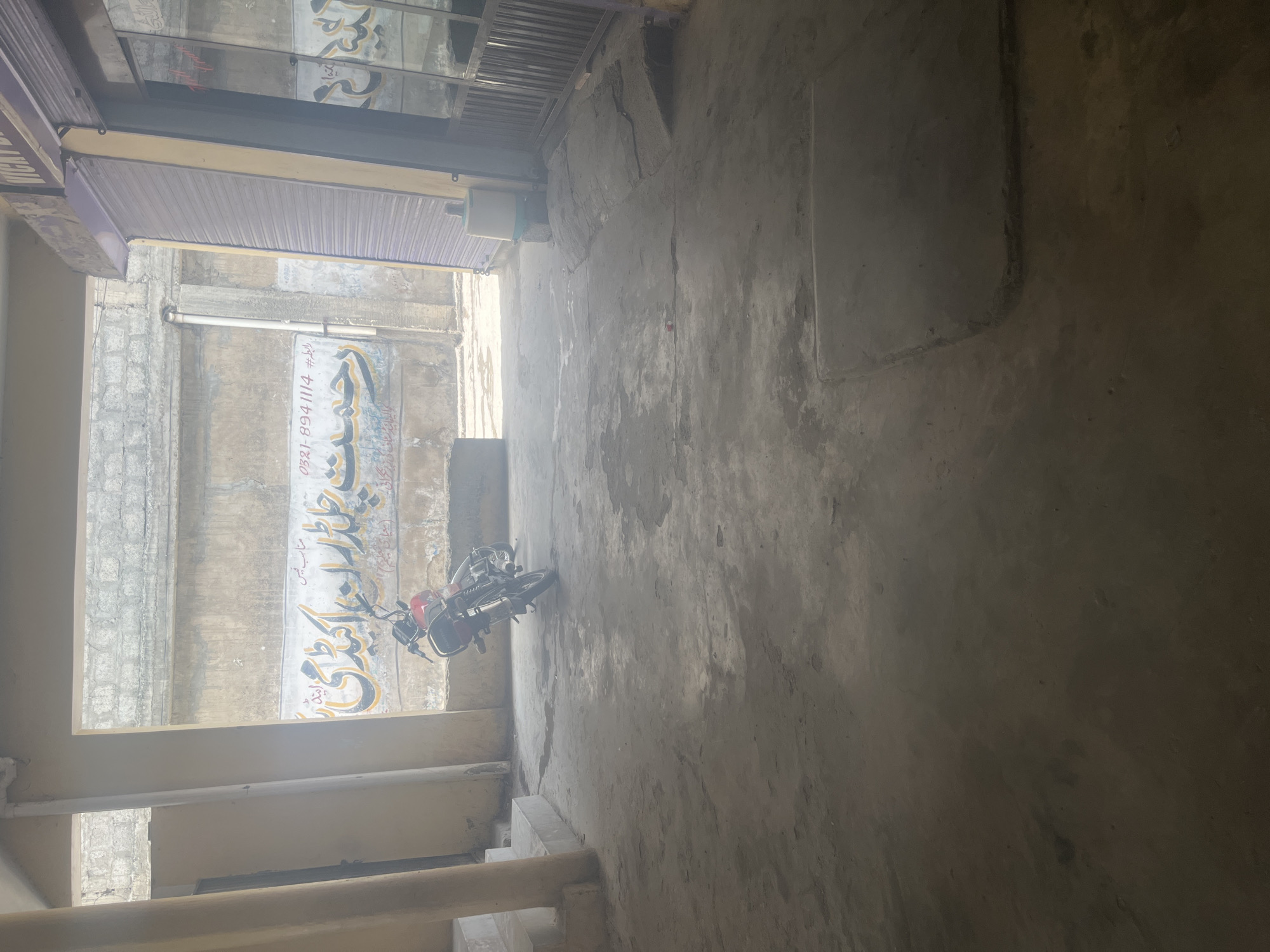 A red motorcycle is parked on a dusty, concrete ground. Above the motorcycle, a mural is painted on a wall. The mural contains Arabic text, with some words written in yellow and others in black. The text appears to be a business name or advertisement. The surrounding environment suggests an outdoor, urban setting, possibly an alleyway or a courtyard. The walls are rough and weathered, and the ground is uneven. Sunlight streams into the area, creating shadows and highlighting the dust.