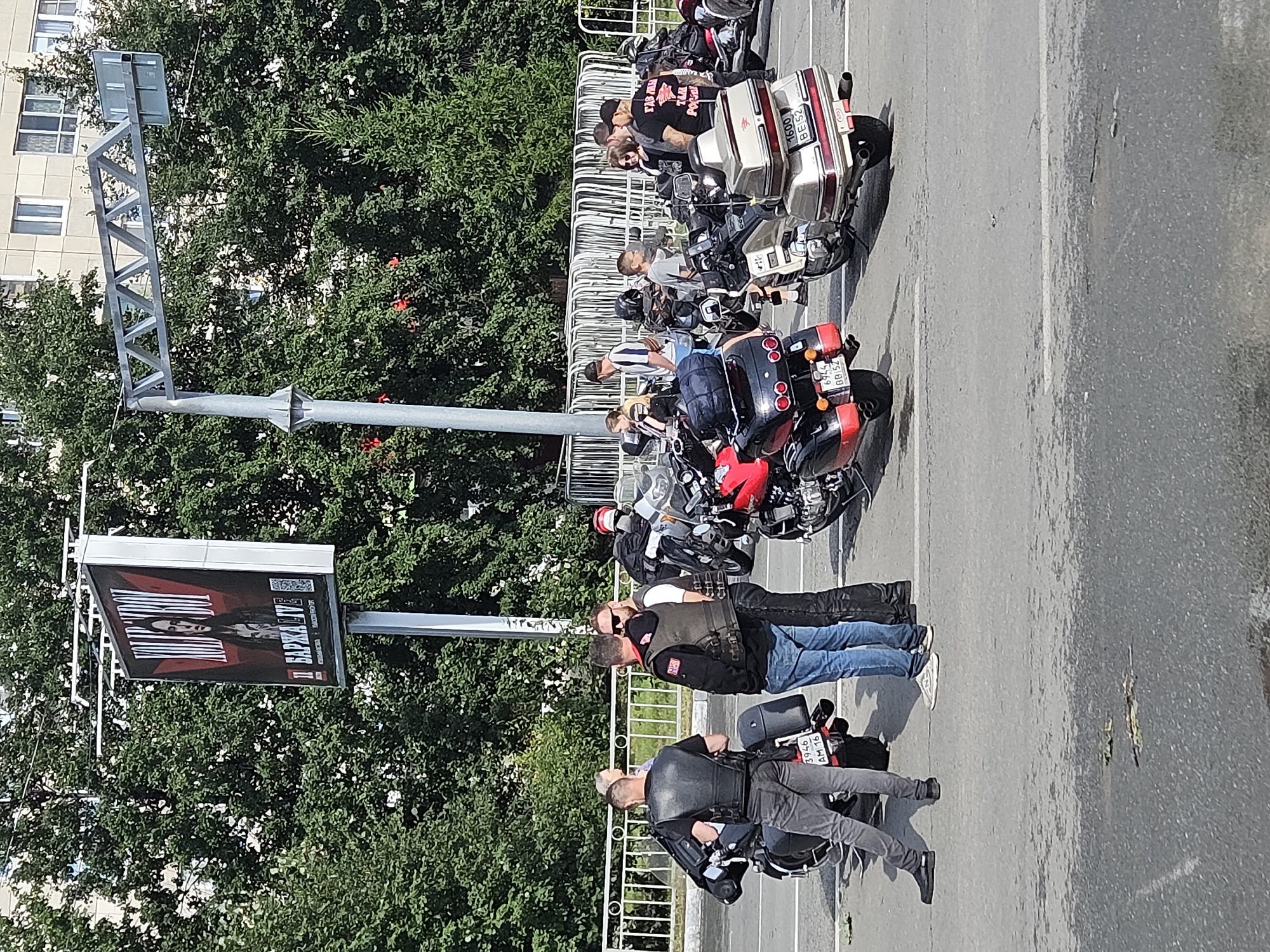 The image captures a scene on a sunny day, likely in the city of Ulyanovsk, Russia, featuring a group of people gathered around several motorcycles. In the background, a billboard displays an image of a person with text in Russian, partially obscured by trees and a metal structure overhead.
The foreground is dominated by a paved road. Several motorcycles are parked, including a black one with a license plate reading "3946 AM 16," a red and black one with license plate "6944 BB152," and a silver one with license plate "1600 BE152."
A group of individuals, mostly men, are seen interacting. Some are wearing black vests, common attire for motorcycle clubs. One man in a black vest and grey pants is standing next to the black motorcycle. Another man in a similar vest but with a red patch and blue jeans is talking to a third man. A few other individuals are visible further back, also near motorcycles. The atmosphere appears casual, with people engaged in conversation.
The environment includes trees lining the background, with a building visible behind them. A white metal fence runs along the edge of the road, separating it from the greenery. The sky is clear and blue. The overall impression is of a gathering of motorcyclists in an urban setting.