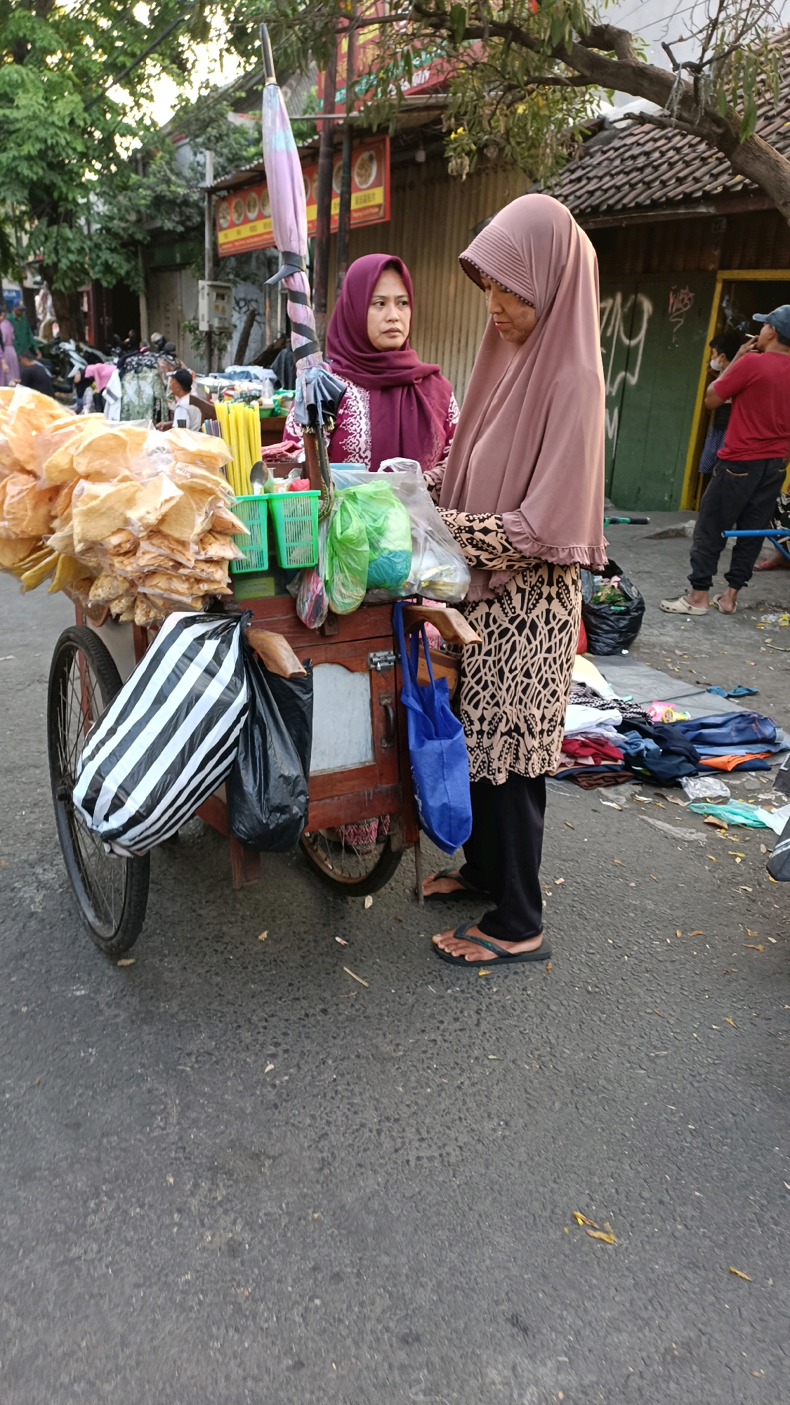 Two women, dressed in hijabs and modest clothing, stand beside a wooden food cart on a paved street. The cart is laden with snacks, possibly crackers, stacked in plastic bags and containers. A pink and purple umbrella is attached to the cart's frame, partially unfurled. The woman on the left wears a maroon hijab and a patterned top, while the woman on the right is clad in a dusty rose hijab and a patterned long-sleeved shirt. The street is lined with trees and buildings, suggesting an urban setting. In the background, other individuals are visible, including a person in a red shirt and a blue cap who appears to be smoking, and a pile of clothing is spread on the ground to the right of the cart. The overall scene depicts a bustling street market environment, likely in Surabaya, Indonesia, given the context of the user's request.