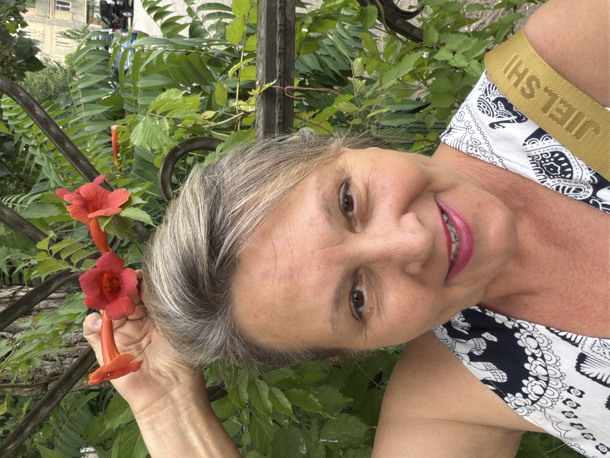 A close-up, selfie-style portrait of a fair-skinned, middle-aged woman with graying blonde hair, taken outdoors. She is smiling with her mouth slightly open, revealing her teeth. She has bright pink lipstick on. Her right arm is raised, and she is holding a cluster of red-orange flowers. She is wearing a black and white patterned top with a yellow strap across her shoulder that has the word "JIELSHI" imprinted on it. The background consists of green foliage from a tree or bush, and a metal fence. Beyond the fence, a street with cars and buildings is visible, suggesting an urban setting. The lighting suggests it is daytime.
