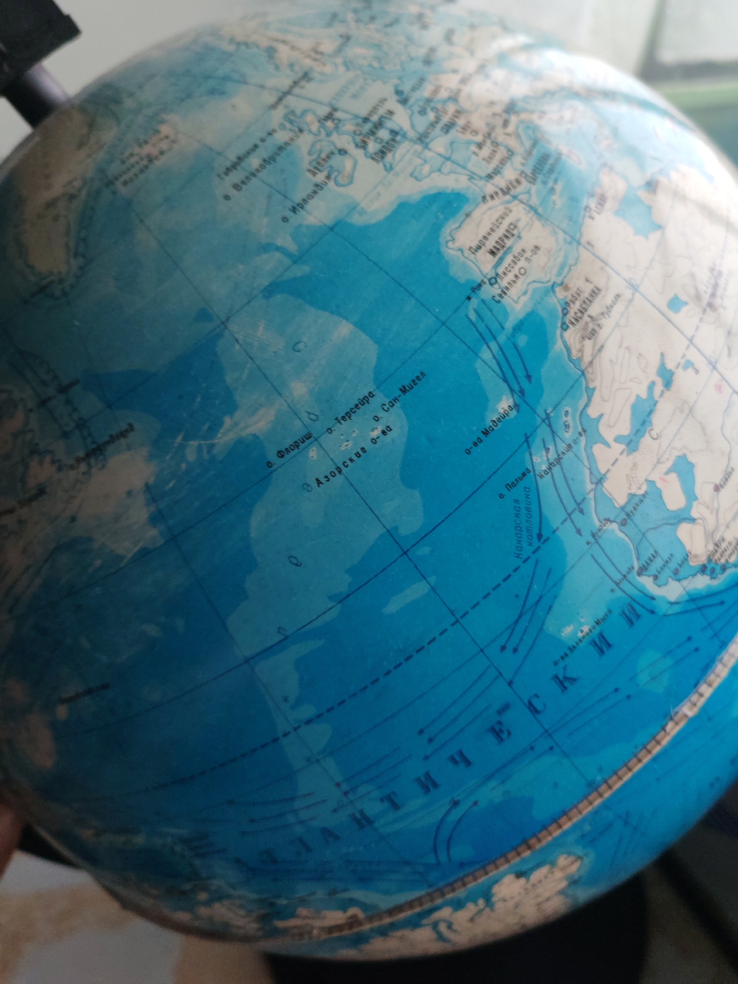 The image is a close-up, slightly blurred photograph of a terrestrial globe. The globe displays continents and oceans with geographical names written in Russian. The focus is on the Atlantic Ocean, with parts of Europe and Africa visible. Several islands are labeled, including the Azores and Madeira. There are also lines indicating ocean currents. The lighting suggests an indoor setting, possibly with natural light coming from the side. The image does not depict the city of Chulman, Russia, nor does it contain any people or specific objects beyond the globe itself.
