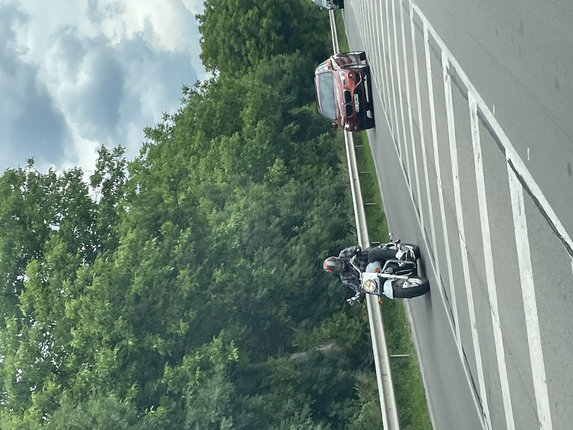 A roadside scene in the city of Getsovo, Bulgaria. A person on a white motorcycle rides on the right side of a multi-lane highway. A maroon BMW car travels ahead. The highway features painted white stripes separating the lanes and a gray metal barrier on the right side of the motorcycle. The background consists of a dense forest. The sky is overcast.