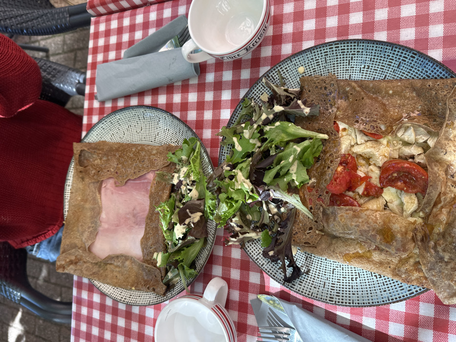 Two savory crepes (galettes) with accompanying side salads are displayed on a red and white checkered tablecloth in an outdoor dining setting in Orléans, France. The upper plate presents a square-folded buckwheat crepe containing a prominent slice of ham, beside which lies a portion of mixed green salad with a creamy dressing. The lower plate features a similar buckwheat crepe filled with chopped white meat, halved cherry tomatoes, and possibly artichoke hearts, also accompanied by a mixed green salad with dressing. Both plates are dark blue and white speckled ceramic. An empty white ceramic cup with red horizontal stripes rests to the left of the upper plate. Another empty white ceramic cup, with a red rim partially inscribed with "fore Kerisac," is situated to the right of the lower plate. A light blue-gray folded paper napkin, concealing cutlery, is placed on the upper right of the table, and another similar napkin with visible cutlery is on the lower left. A person wearing a red knitted garment and blue jeans is partially visible in the background, seated on a dark, woven-style chair behind the upper plate. The arrangement indicates a meal prepared for at least two individuals.