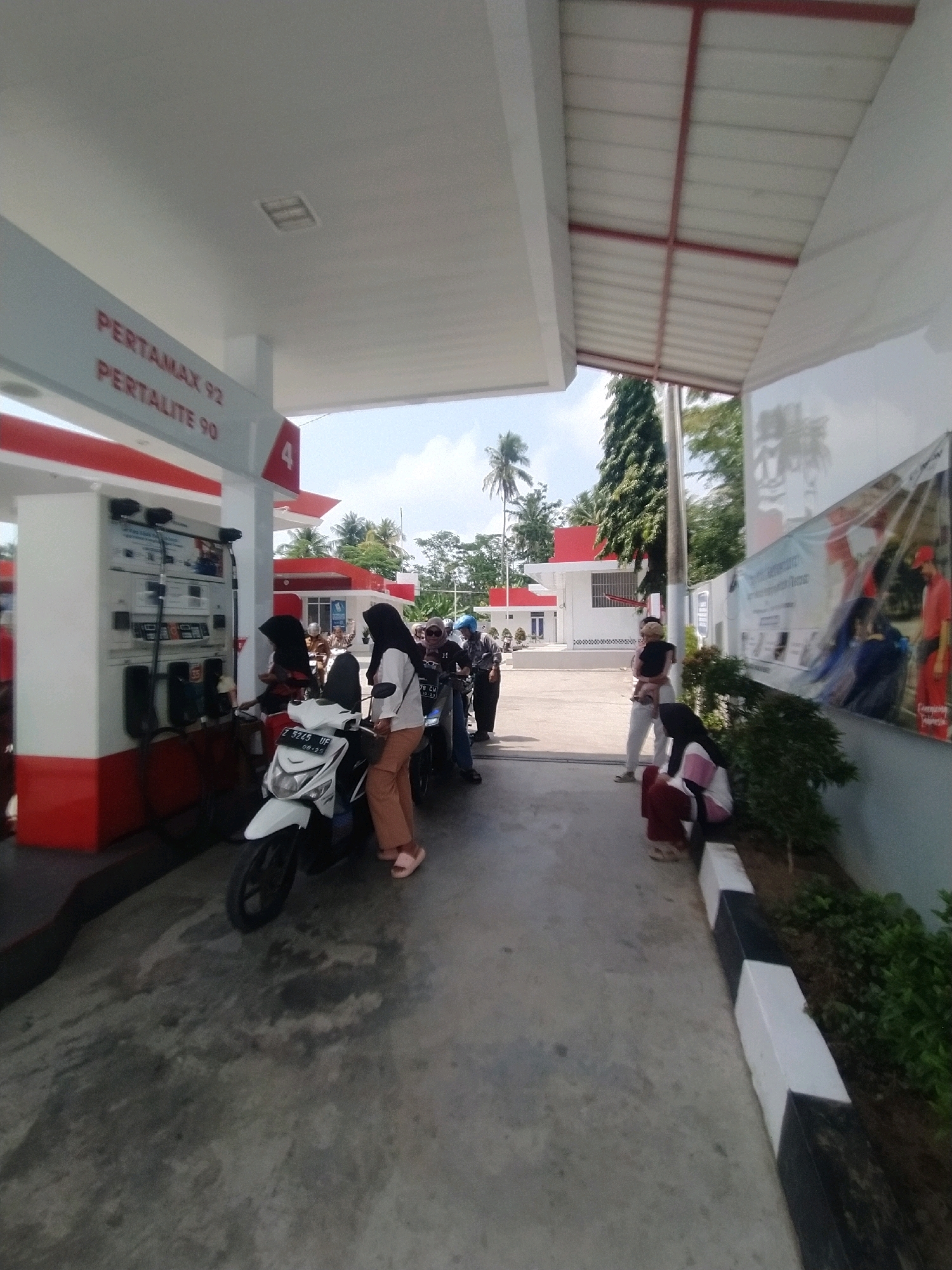A gas station with a white and red canopy is shown. On the left, a gas pump is visible with a sign that says "PERTAMAX 92" and "PERTALITE 90." Several people on motorcycles are waiting to refuel. A white motorcycle is in the foreground, and to its right, a woman in a hijab and brown pants is standing. Further back, a few more people and motorcycles are visible. In the background, a building with a red roof and white walls is seen, along with palm trees and other vegetation. The ground is paved. The scene appears to be in a rural or semi-urban area.