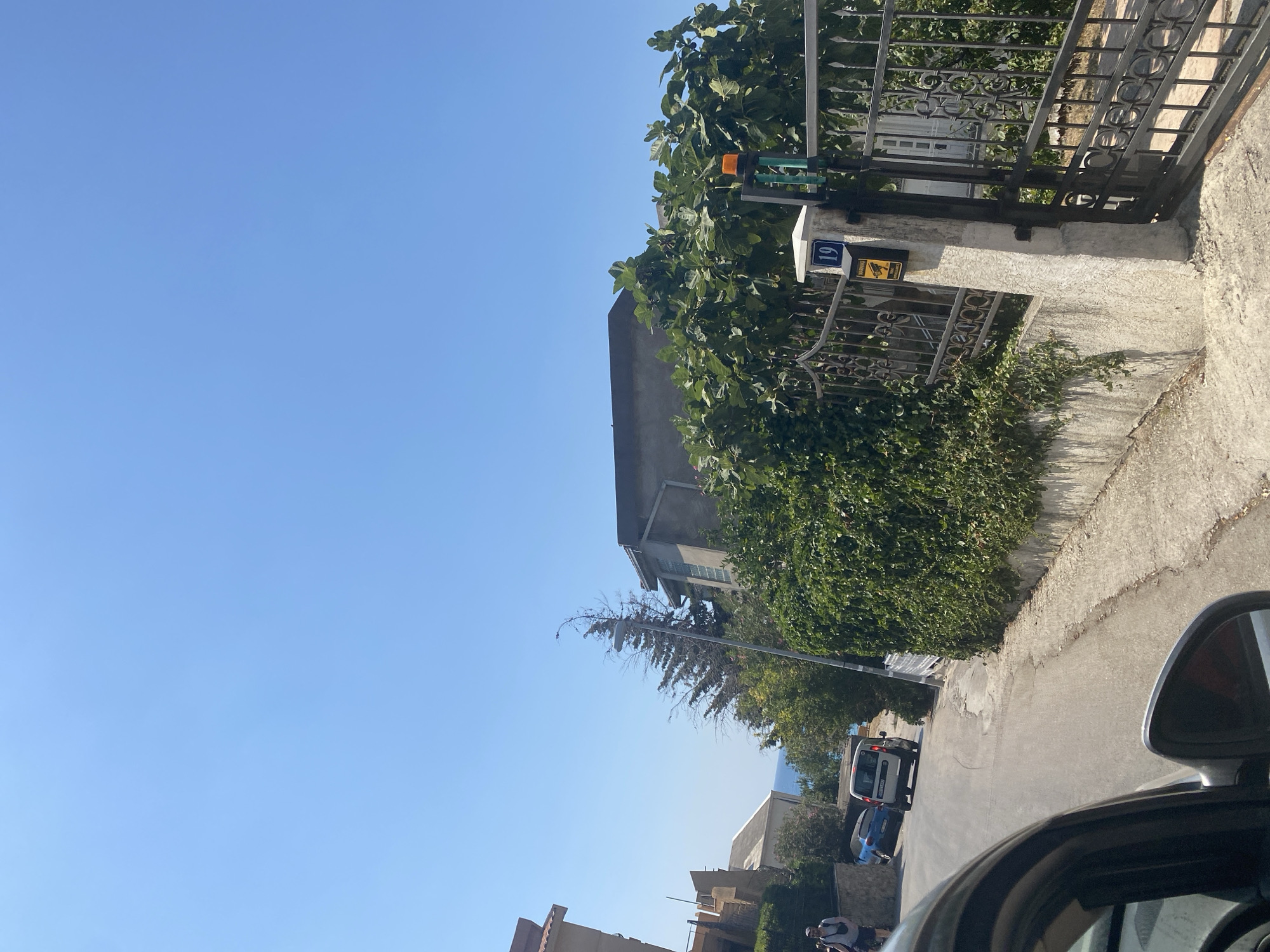 The image depicts a sunlit street scene in the city of Bar, Montenegro, likely in a residential area, possibly on a hillside given the incline of the road. The sky is a clear, bright blue, suggesting a warm, sunny day. In the foreground, a portion of a dark-colored car's interior is visible, including the side mirror. The car is positioned on a paved road that curves upwards.
Further up the street, a white van and a blue car are parked. On the left side of the road, several buildings are visible, some appearing residential with balconies and stone facades. Greenery, including bushes and trees, is abundant, lining the street and climbing over some walls.
On the right side of the road, a black metal gate stands open, leading to a property. A concrete pillar next to the gate is topped with a blue sign displaying the number "19". A yellow warning sign with a camera icon is affixed to this pillar. An orange flashing light is mounted on top of a green pole near the gate. Behind the gate, dense green foliage, possibly a fig tree with visible figs, dominates the scene, with glimpses of a building structure behind it. The overall impression is one of a quiet, residential street bathed in bright sunlight.