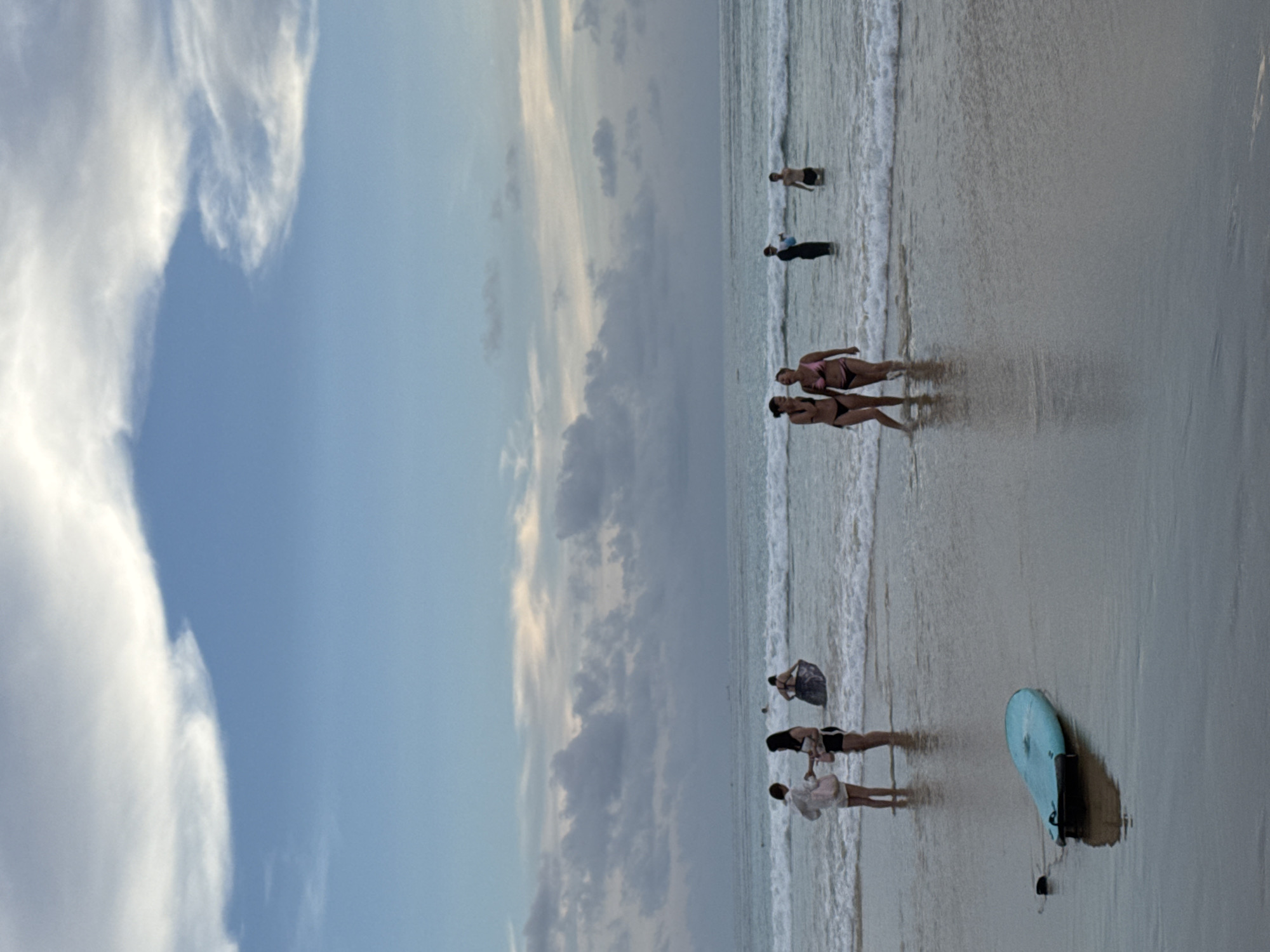 The image depicts a beach scene in Karon, Thailand, during a cloudy dusk. In the foreground, a light blue surfboard rests on the wet sand. The middle ground shows several people wading in the shallow water. Two women in bikinis are walking towards the viewer, their upper bodies visible, with one woman laughing. Further back, two individuals stand in the water, one holding a blue object. In the background, a person in the distance is partially submerged in the water. The ocean exhibits moderate waves, and the sky is characterized by a mix of blue sky and dramatic, overcast clouds.
