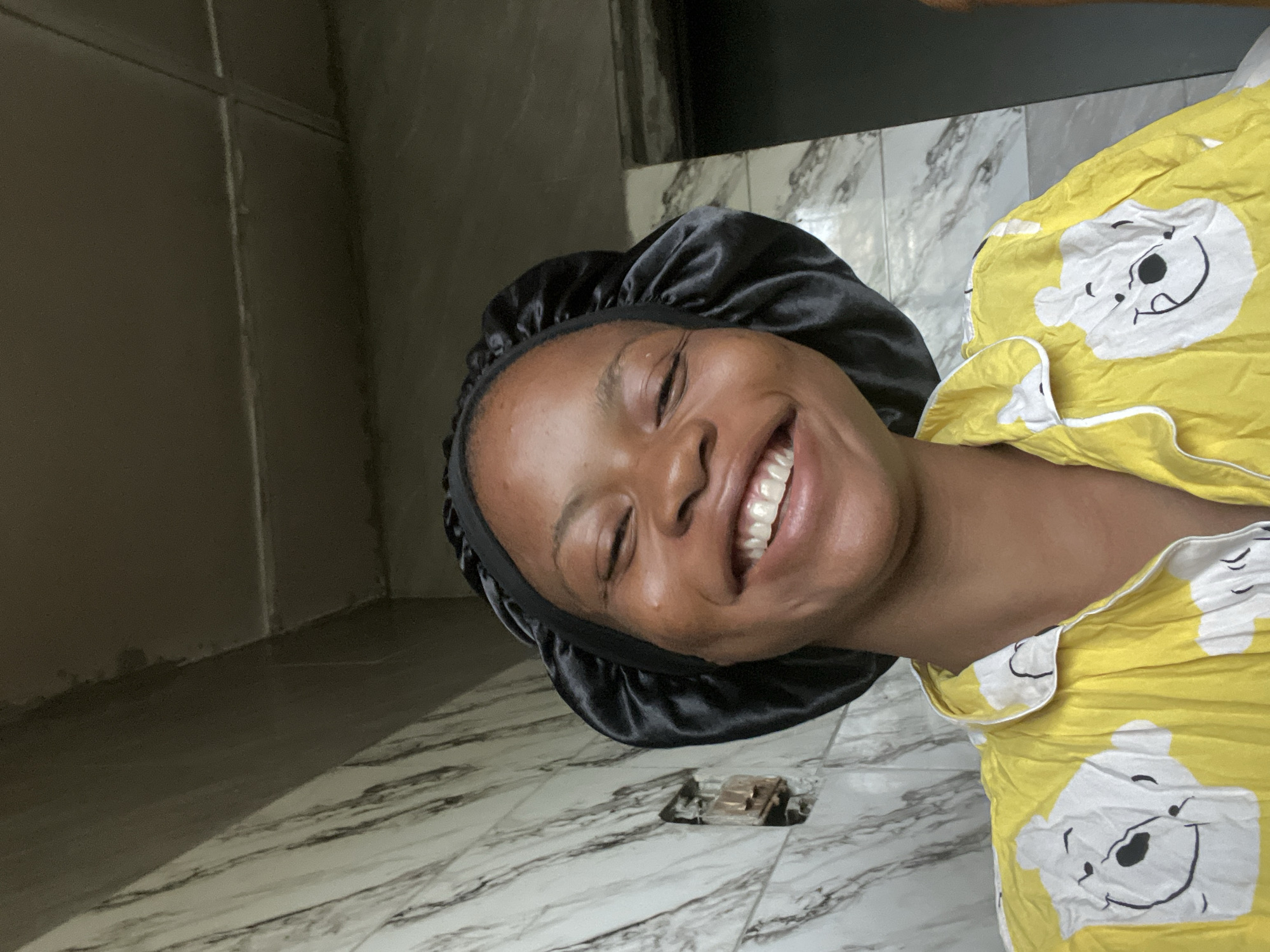 A young Black woman is depicted in a close-up, indoor portrait, smiling broadly with her eyes closed, suggesting happiness and amusement. Her head is tilted to the right, and she is wearing a shiny black satin bonnet. She is dressed in a vibrant yellow pajama top adorned with a white pattern featuring Winnie the Pooh characters.
The background consists of a dark, possibly grey or brown, wall or piece of furniture in the upper left. The dominant surface is a white and grey marble-patterned tiled floor, which covers the lower and right portions of the image. A small, metallic, recessed object, possibly a floor outlet or fixture, is visible within the tiles below her chin. The setting appears to be a room, with soft, even lighting. The image conveys a cheerful and lighthearted mood. The location context for this scene is Lagos, Nigeria.
