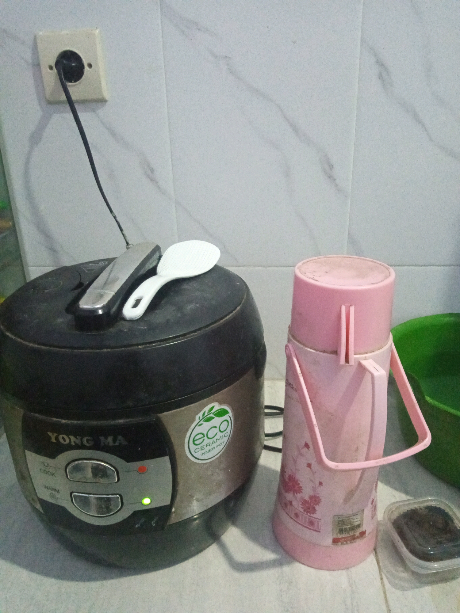 In Sumbergirang, Indonesia, a rice cooker with a "YONG MA" logo sits on a tiled surface. A white rice paddle rests on top of the rice cooker. Beside the rice cooker is a pink thermos. A power outlet with a black cord is visible on the tiled wall behind the appliances. A small clear container with dark contents is to the right of the thermos.