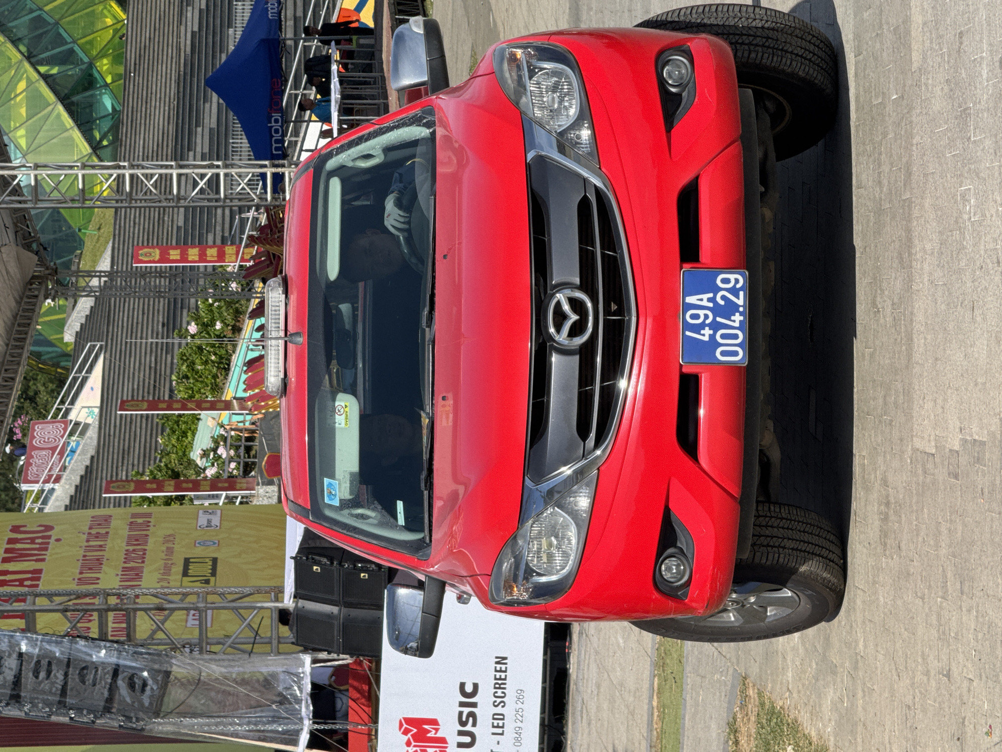 A bright red Mazda BT-50 pickup truck is parked on a paved area, with a grassy verge to the left. The truck is facing the camera and has its headlights on. The license plate reads "49A 004.29". Inside the truck, a person wearing a blue shirt and white gloves is visible behind the steering wheel. A light bar is mounted on the roof of the truck. In the background, there is a large stage with a yellow banner displaying Vietnamese text. Behind the stage, there are tiered seating areas and a building with a green, geometric facade. A blue Mobifone tent is also visible to the right of the stage. The overall scene appears to be an outdoor event or festival. The license plate suggests the vehicle is registered in Da Lat, Vietnam, as "49" is the provincial code for Lam Dong province, where Da Lat is located.