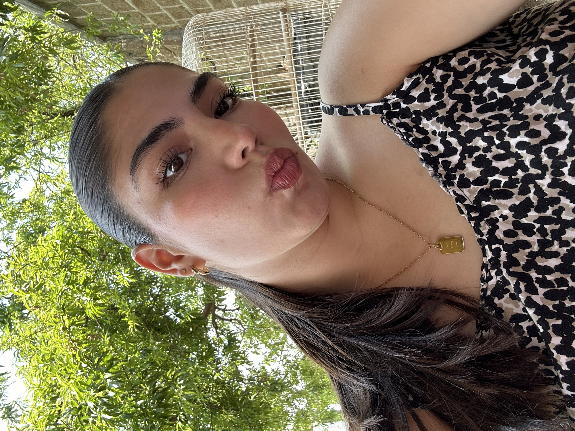 A woman with dark hair, styled in a sleek ponytail, and wearing a black and white patterned top is positioned in front of a leafy green tree. The location appears to be outdoors, likely in a garden or park setting. A wire structure can be seen in the background. The woman is looking directly at the camera with a slight pout. She is wearing a gold necklace with a rectangular pendant and earrings.