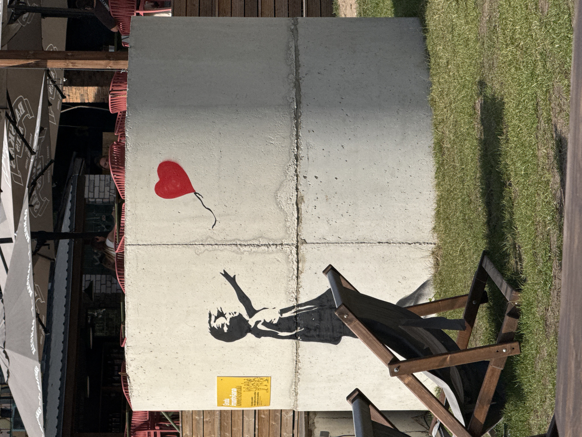 The image depicts a concrete cylinder with graffiti. On the left side of the cylinder, a stencil of a child is depicted reaching out towards a red heart balloon that appears to be floating away. A yellow sign with text in Latvian is posted to the left of the stencil. The concrete cylinder is situated outdoors, with a grassy area in the foreground where two wooden deck chairs are placed. In the background, there are umbrellas, tables, and chairs, suggesting an outdoor seating area for a cafe or restaurant. The overall scene appears to be in an urban environment, possibly a public space or a recreational area. The style of the graffiti is reminiscent of Banksy's work. While the specific location in Riga, Latvia is not explicitly stated in the image itself, such street art installations are often found in various public areas within cities.