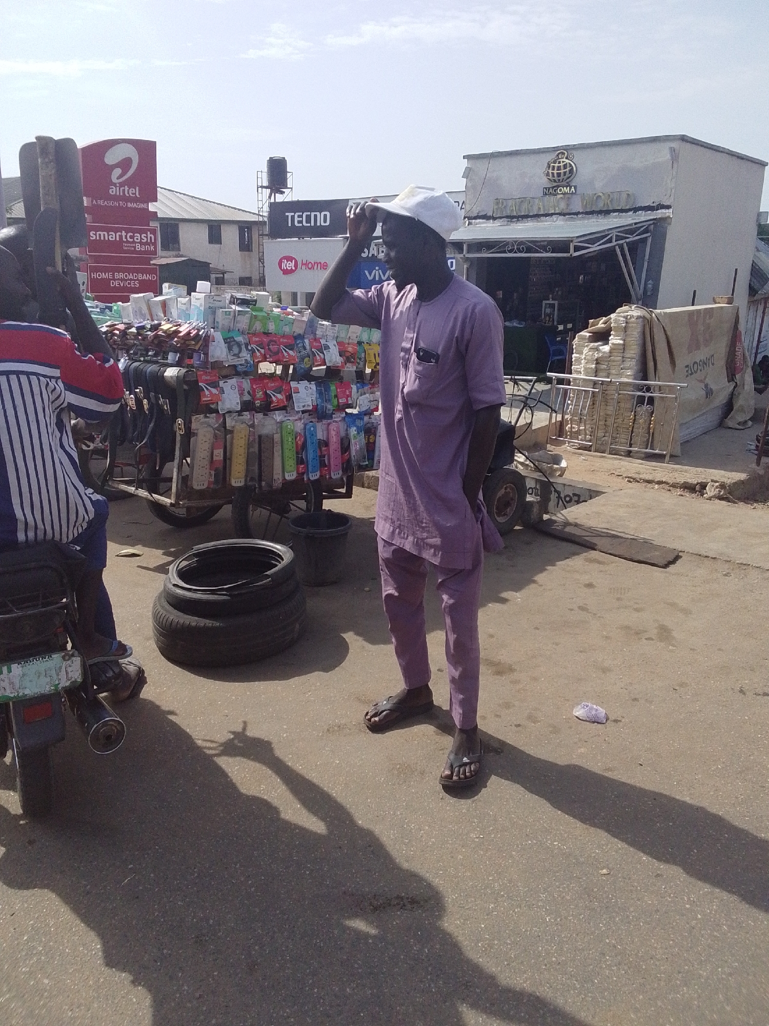 The image captures a street scene in Lokoja, Nigeria, under a bright sky. The main subject is a man dressed in a lavender-colored, short-sleeved traditional outfit and a white cap, with his hand on his head. He is standing on a paved street, casting a long shadow. In the background, there is a roadside vendor with a cart laden with items, possibly phone accessories. Signs for Airtel, Smartcash Bank, Tecno, and Itel Home are visible above the cart. To the right, there is a building with the name "Nagoma" on it. A motorcycle with a rider is to the left of the frame. A stack of used tires rests on the ground near the vendor's cart. A white plastic bag lies on the pavement.