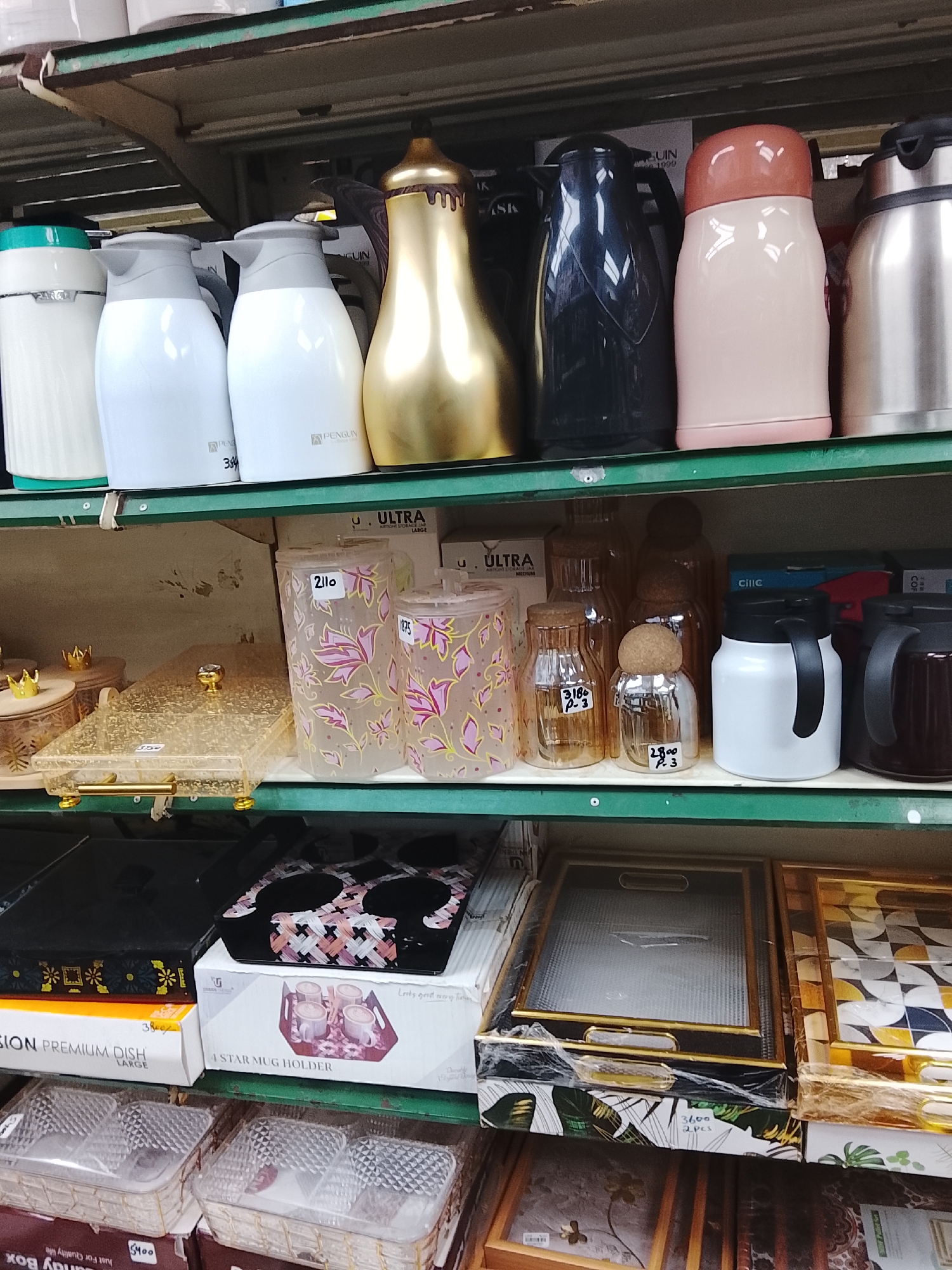 The image shows a shelf stocked with various household items in a retail setting, likely in Gulgasht Colony, Pakistan. The top shelf displays insulated beverage containers in a range of colors and styles, including white, gold, black, and pink, with brands such as Penguin visible. The middle shelf features plastic containers with floral designs, glass jars with cork stoppers, and white and black mugs. Below, the bottom shelf holds trays and boxes, including a box labeled "4 STAR MUG HOLDER" and "SION PREMIUM DISH LARGE." Price tags are visible on several items, indicating the costs in Pakistani Rupees. The setting suggests a store or market specializing in household goods.