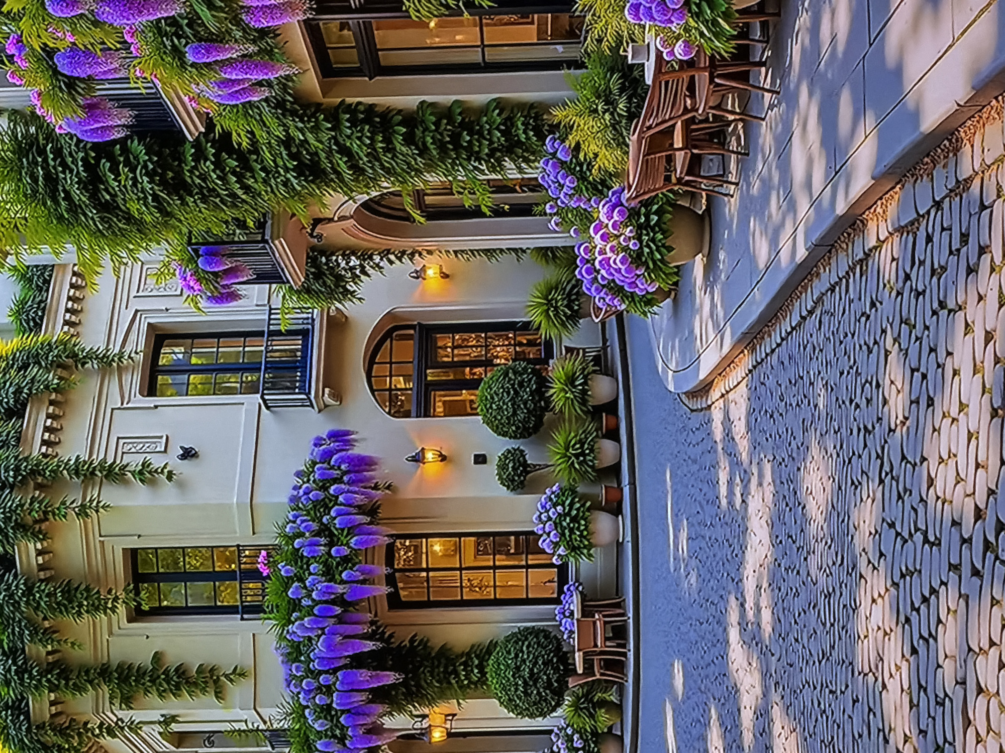 This image depicts a charming and elegantly adorned building facade in Sofiivska Borshchahivka, Ukraine, viewed from a cobblestone path. The building features a light-colored (cream or off-white) exterior with dark-framed windows and doors, some of which are arched, giving it a classic architectural feel. Small ornate railings are visible on some windows.
The facade is extensively covered with lush green climbing plants and adorned with abundant clusters of vibrant purple flowers, possibly wisteria or similar cascading blooms, creating a striking visual contrast. Additional purple flowers are arranged in large decorative planters at ground level, alongside neatly trimmed green bushes. Wall-mounted sconces cast a warm, inviting glow near the doorways.
On the right side of the image, a paved cobblestone path or street is visible, with long, soft shadows stretching across it, suggesting either early morning or late afternoon sunlight. Several empty wooden chairs are placed on what appears to be an outdoor seating area or terrace, hinting at a cafe or an inviting spot for relaxation.
No people are present in the scene, contributing to a tranquil and serene atmosphere. The overall impression is one of refined beauty and natural abundance. No discernible text is visible within the image.
