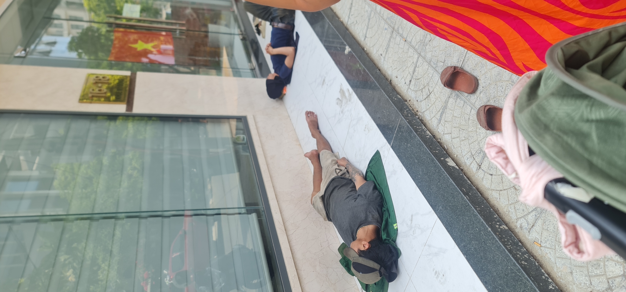 A high-angle shot depicts a scene outside a building in Đà Nẵng, Vietnam. The building's reflective facade features a Vietnamese flag and a sign with the number 18. Two men are lying on the ground next to the building. One man, wearing shorts and a t-shirt, is lying on a green mat. The other man is wearing a blue shirt and dark pants. Next to them is a pair of brown sandals and a multi-colored blanket. A green bag and a pink shirt are also visible in the foreground.