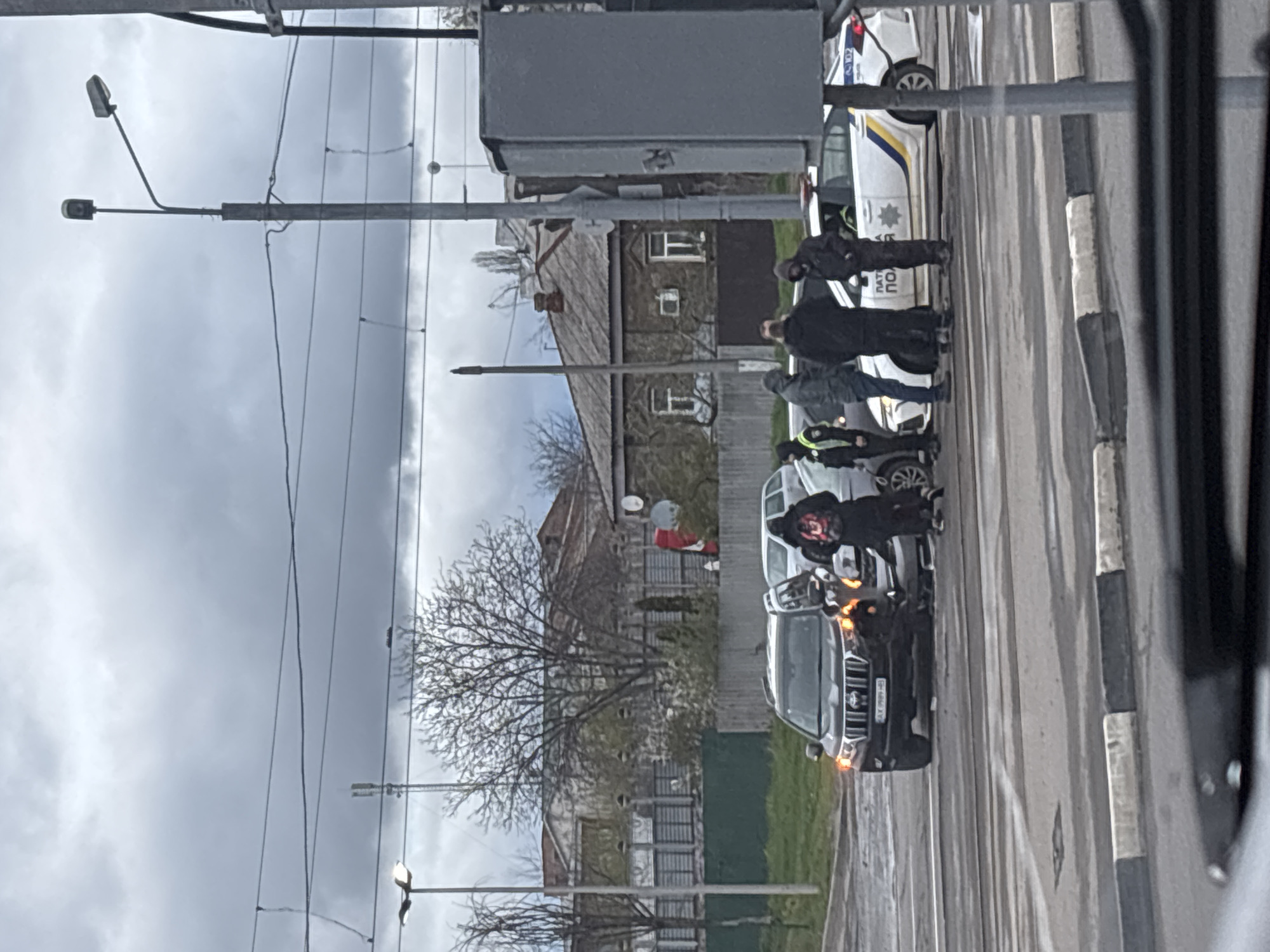 The image captures an outdoor street scene in Kharkiv, Ukraine, on an overcast day. The original image is rotated 90 degrees counter-clockwise; when viewed correctly, the horizon is level.
Several individuals are gathered on what appears to be a street or intersection. Two white police cars, marked with "ПАТРУЛЬНА ПОЛІЦІЯ" (Patrol Police) and an "02" emergency number, are parked on the right side of the road. One police officer is visible standing near a police car. A dark-colored SUV, possibly a Toyota Highlander, is in the foreground with its hazard lights illuminated, suggesting an incident. Its license plate "XA 9999 HY" is partially visible. Another light-colored car is positioned behind the SUV.
Among the people, one individual near the SUV wears a dark hoodie with a distinctive red and black pattern. Another person in the mid-ground stands out in a white protective suit with red elements on the lower body, indicative of a potential hazardous materials or medical situation.
The background features old, multi-story stone or brick buildings with windows, typical of an urban or suburban environment. Bare trees are visible, suggesting late winter or early spring. Overhead, numerous power lines crisscross the grey sky, and streetlights are mounted on utility poles. A large grey utility box is also part of the urban infrastructure. The presence of multiple police vehicles and a person in a protective suit indicates an ongoing official response to an event, possibly an accident or an incident requiring specialized personnel.
