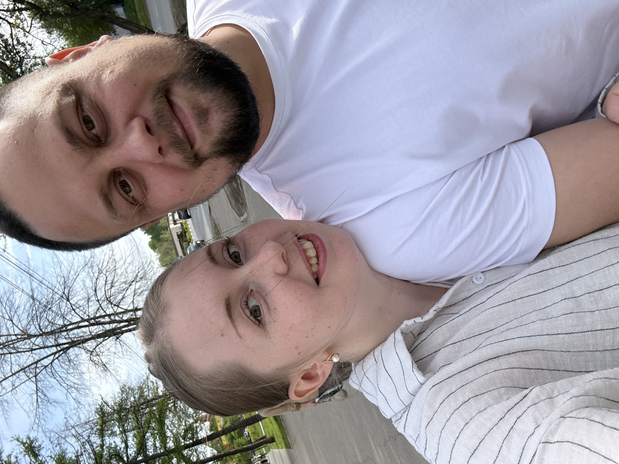 A selfie shows a light-skinned woman and a light-skinned man in an outdoor urban setting, seemingly a residential street.
The woman is wearing a light-colored striped shirt with buttons and a pearl earring. The man, slightly above and behind her, is wearing a white t-shirt. The background features trees with bare branches and some evergreen trees. The pavement of a street is visible, along with some parked vehicles and street infrastructure.