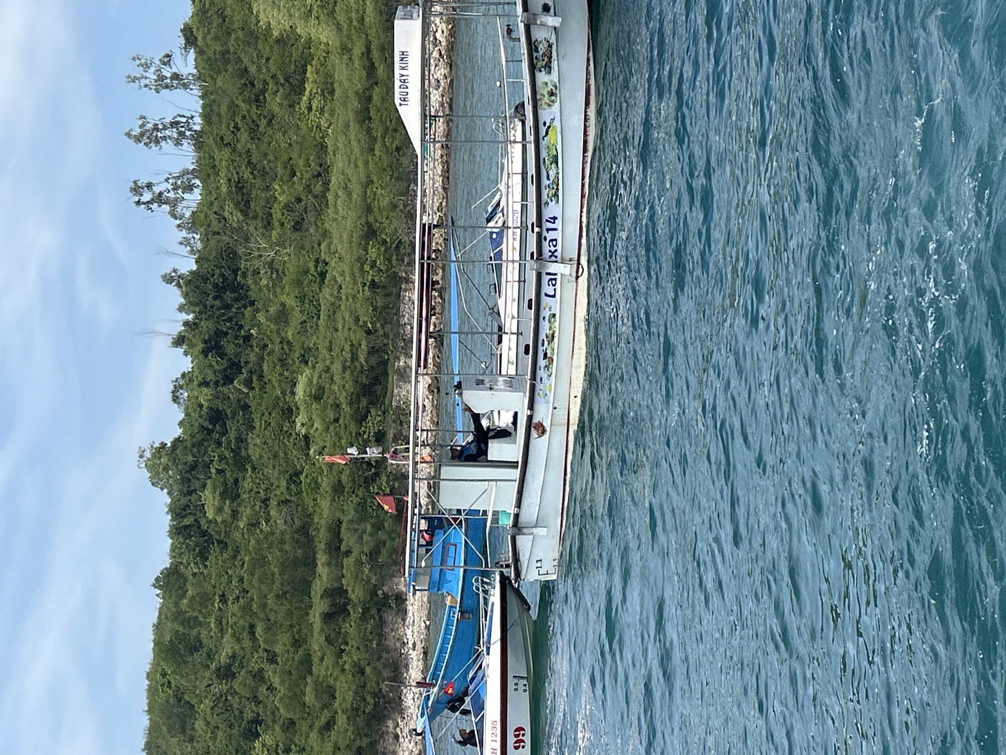 The image depicts a sunny day at sea near a verdant, tree-covered island, likely off the coast of Nha Trang, Vietnam. The water is a clear, vibrant blue, with small waves indicating a gentle sea state. In the foreground, a white tour boat with a covered seating area is prominently featured. The boat has colorful decals of marine life along its hull and the number "13" is visible near the bow. A person in dark clothing is seated on the right side of the boat, with their legs extended. A smaller white boat with the number "99" and registration "H 1235" is moored to the left of the tour boat, with two individuals visible on board. Further in the background, a bright blue boat is also present. Several Vietnamese flags are displayed on poles, particularly on the tour boat. The island's shoreline is visible in the distance, densely covered with lush green vegetation, suggesting a tropical environment. The sky is a bright blue with scattered, wispy clouds. The scene captures a typical tourist activity or transportation scenario in the waters of Nha Trang.