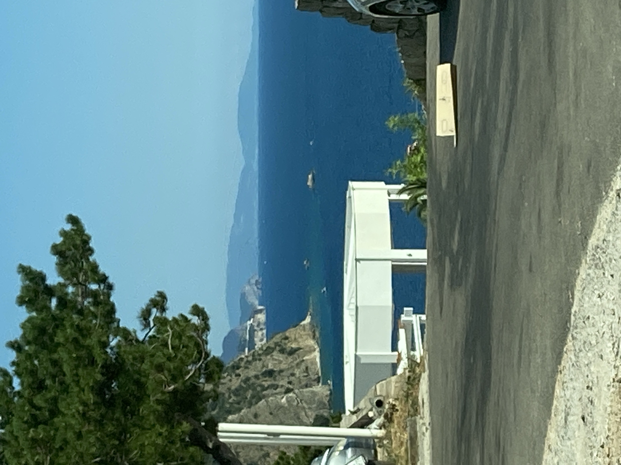 This is a medium-range, eye-level shot of a scenic view of the Adriatic Sea near Budva, Montenegro. In the foreground, a paved road curves towards the right, with gravelly shoulders on either side. A single, light brown, cardboard object, possibly a sign or packaging, rests on the road surface. In the midground, a large, white, octagonal structure with supporting pillars overlooks the sea, partially obscured by a dense evergreen tree on the left. The sea itself is a vibrant blue, dotted with a few small boats in the distance and a rocky outcropping. Steep, rocky cliffs rise from the water on the left, with buildings and vegetation visible along their upper edges. In the far distance, a chain of hazy, blue mountains stretches across the horizon under a clear, light blue sky. The overall impression is one of natural beauty and coastal development.