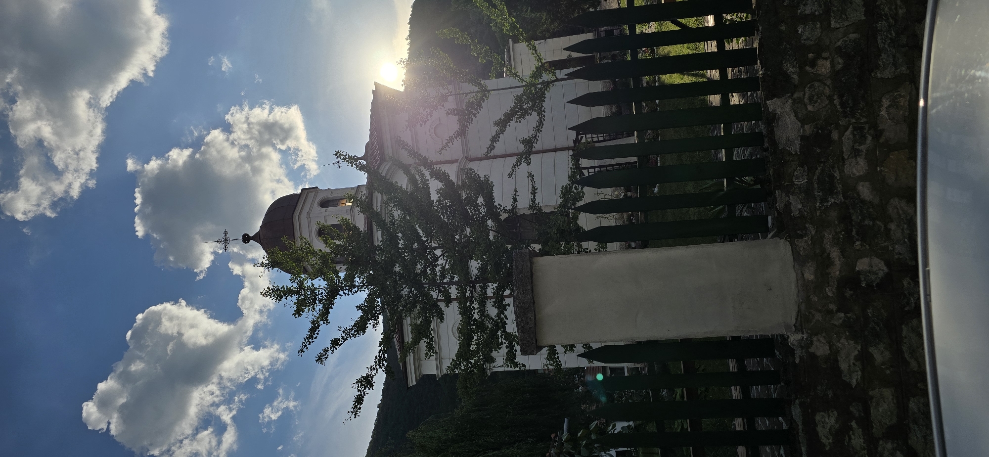 A white church with a dark dome and a cross on top is pictured with a tree in the foreground. The church is located in a hilly, green area. A wooden fence is in front of the church, and a stone wall is at the bottom of the image. The sun is setting behind the church, casting a warm glow. The sky is blue with white clouds.