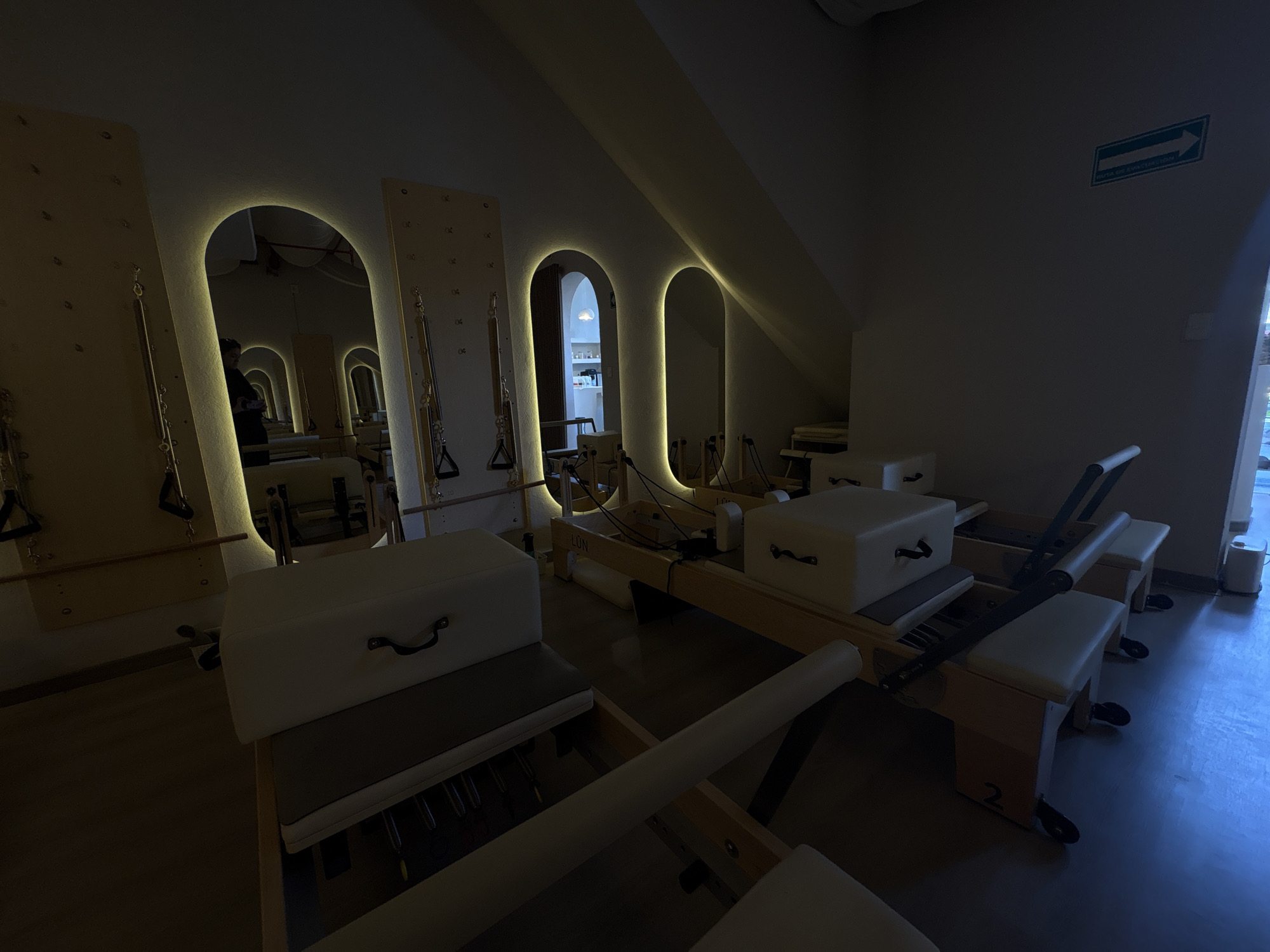 The image shows a dimly lit indoor space, possibly a pilates studio in Mexico City, Mexico, judging by the emergency exit sign. The studio contains several pilates reformer machines arranged in a row. The reformers have wooden frames and white padded cushions, and some have number labels. There are mirrors with arched, lit frames along one wall. The room has an angled ceiling. The overall atmosphere is dark and quiet.