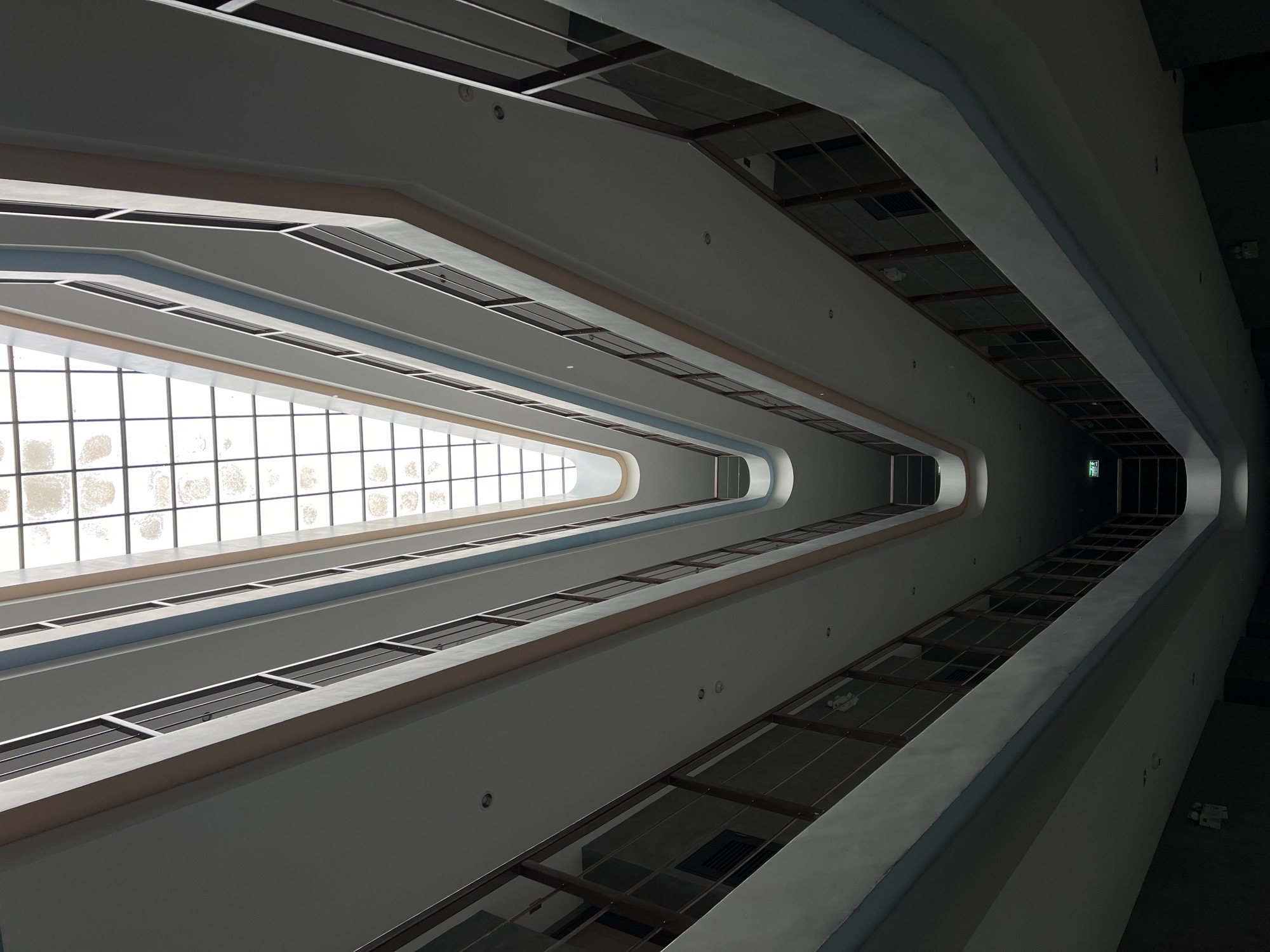 The image depicts a view looking upwards through a multi-story atrium or interior courtyard. The atrium is defined by a series of receding levels, each with a rectangular opening. The sides of each level appear to be painted white or light gray with blue and tan trim. There is a large, rectangular skylight at the top of the atrium, constructed with a grid-like pattern of glass panels. At the other end of the structure there is an exit sign. The walls appear to be featureless except for the trim and a few small light fixtures. There are no people visible in the photograph. The building's location is unknown.