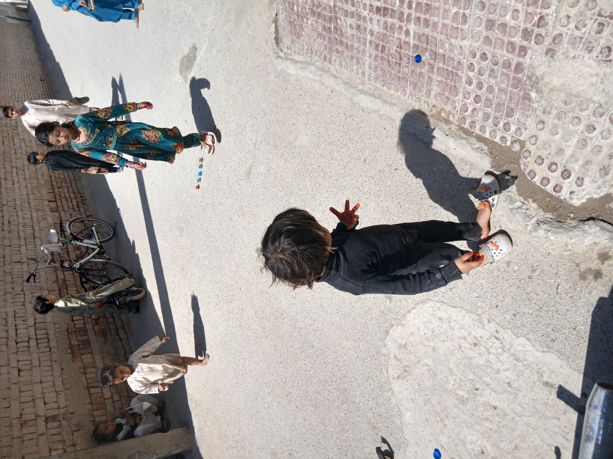 The image depicts an outdoor scene in Quetta City Tehsil, Pakistan. Several individuals are present, including children and adults, engaged in various activities. One child, wearing a black outfit, is seen adjusting their shoe. A girl in a green patterned dress appears to be in motion, possibly playing a game with colored objects. Other children are present, some sitting, some lying near a bicycle, and one standing in light-colored clothing. Several adult figures are visible in the background.
The environment consists of a concrete or paved area alongside a brick wall. A brick wall design is visible on the right side. The lighting suggests it's daytime, casting shadows of the people and objects. A blue bottle cap is visible on the paved surface.