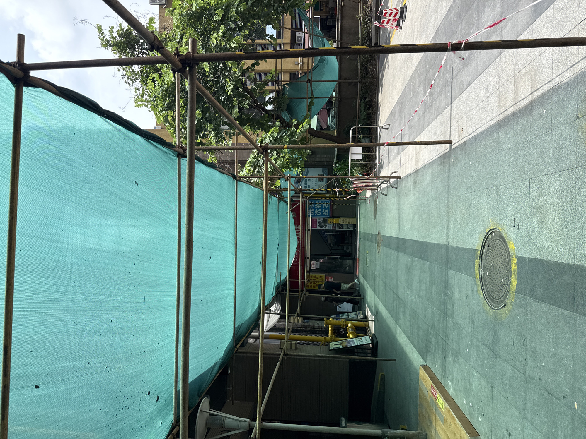 The image shows a covered walkway with a turquoise tarp supported by scaffolding. The walkway appears to be in a city, possibly in Haizhu District, China, given the presence of Chinese text on signs and the architectural style. The ground is paved with large tiles, and there are yellow markings around a manhole cover. Barriers and construction cones suggest ongoing work or restricted access. A few individuals are visible in the distance, walking towards the entrance of a building. Trees and other buildings are visible in the background, indicating an urban environment. The lighting suggests it is daytime, with sunlight casting shadows.