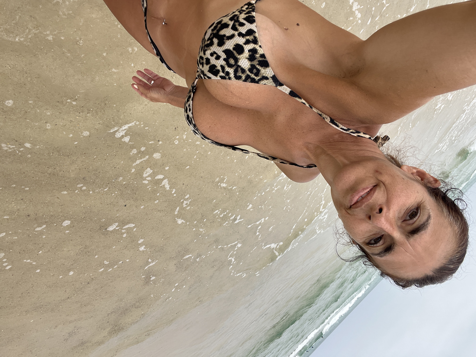 The image is a selfie taken on a sandy beach with the ocean in the background. A woman with brown hair and a leopard print bikini is looking at the camera. She is standing in shallow water with waves gently lapping at the shore. The sky is overcast. The image does not contain any information related to the city of Caldas da Rainha, Portugal.