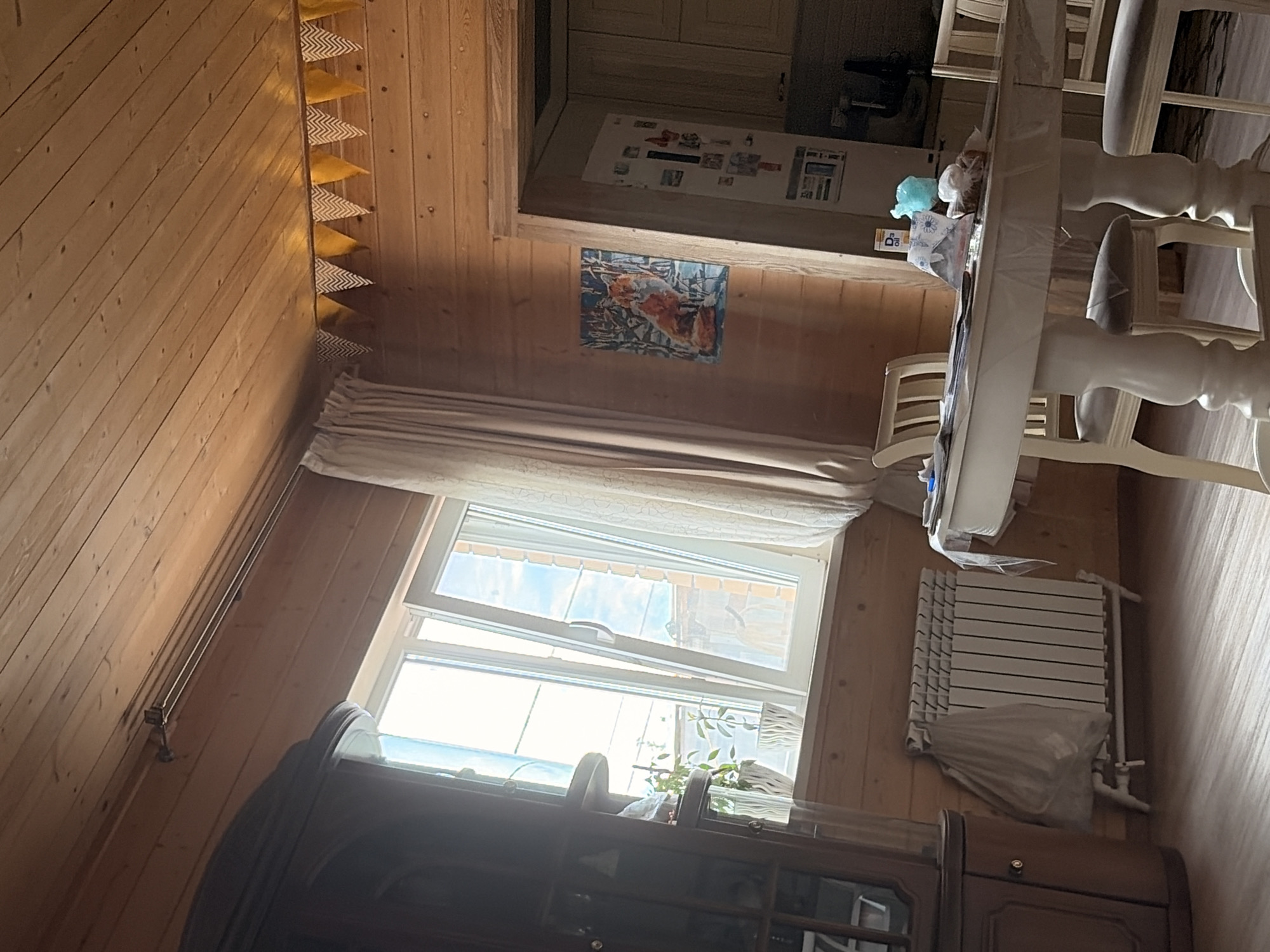 The image depicts the interior of a room with wooden paneled walls and ceiling, likely a residential space in Leninogorsk, Russia. A large window with white frames is visible on the left, letting in bright daylight and showing a glimpse of the sky and possibly outdoor structures. In front of the window, there is a white radiator covered with a translucent plastic bag. To the left of the window, a tall, dark wooden cabinet or hutch is partially visible.
In the center of the room, a round white dining table with carved legs is set with several chairs. The table surface has some items on it, including what appears to be a plastic tablecloth, papers, and a few small objects. To the right of the table, a doorway leads to another area, likely a kitchen, where a refrigerator with magnets on its door and some cabinetry are visible.
On the wooden wall to the right of the window, a framed picture of a fox sitting in a snowy landscape is hung. Further up the wall, near the ceiling, a string of triangular yellow and white pennant banners is displayed. The floor is made of light-colored wood planks. The overall impression is of a lived-in, rustic interior. There are no people visible in the image.