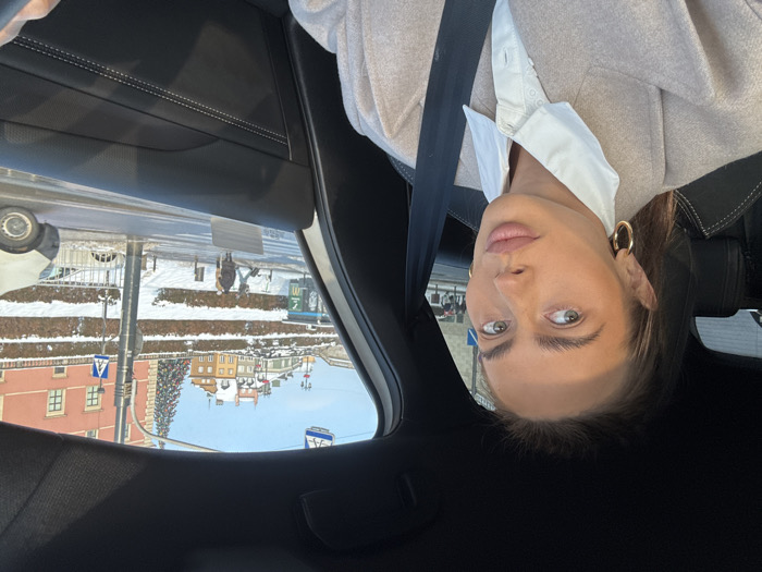 A selfie-style photograph captures a young adult female seated in the driver's position of a vehicle during winter daylight. The subject, a light-skinned woman with light brown hair pulled back, light-colored eyes, and prominent eyebrows, wears a white collared shirt, a beige overcoat, and gold hoop earrings. She is secured by a black seatbelt across her chest and shoulder and looks slightly to her right, away from the camera. Direct sunlight illuminates the left side of her face. The car's interior features black leather upholstery with white stitching on the door panel and seat.
The background outside the vehicle window depicts a snowy urban landscape in Warsaw, Poland. This scene is identifiable as the Plac Zamkowy (Castle Square) area, likely viewed from Krakowskie Przedmieście. An elevated embankment, covered in snow and bare shrubs, separates the street from a row of historical buildings. To the right, a prominent red-brick building, part of the Royal Castle or adjacent structures, is visible. Further left on the elevated ground, several colorful buildings characteristic of Warsaw's Old Town, with snow-covered roofs, are seen. A tall, decorated Christmas tree with red ornaments stands near the buildings to the right. The street in the mid-ground features a pedestrian crosswalk. Several pedestrians are visible on the snowy sidewalk and street, including one individual on a scooter. A light-colored van is partially visible on the right side of the street. A blue bus shelter or advertising panel with a partial yellow McDonald's logo is situated on the left side of the street. Traffic signage, including a blue pedestrian crossing sign, is also present. The sky is clear and bright.