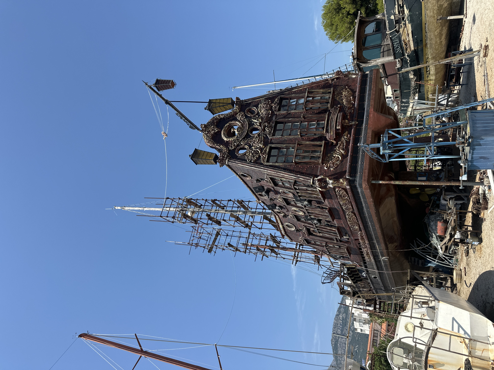 The image shows a large, elaborately decorated ship, resembling a pirate ship, on dry land, possibly in a shipyard or docked for repairs. It is surrounded by scaffolding and other boats. The ship is made of dark wood with ornate golden decorations. It has multiple windows, lanterns, and masts. To the left, a smaller white boat is visible. To the right, another boat with the name "SONJA" is partially visible. The background shows buildings and mountains, suggesting an urban or coastal setting. The sky is clear and blue. The overall impression is one of a historical or fantasy-themed vessel undergoing maintenance or restoration. The location is identified as Bar, Montenegro.