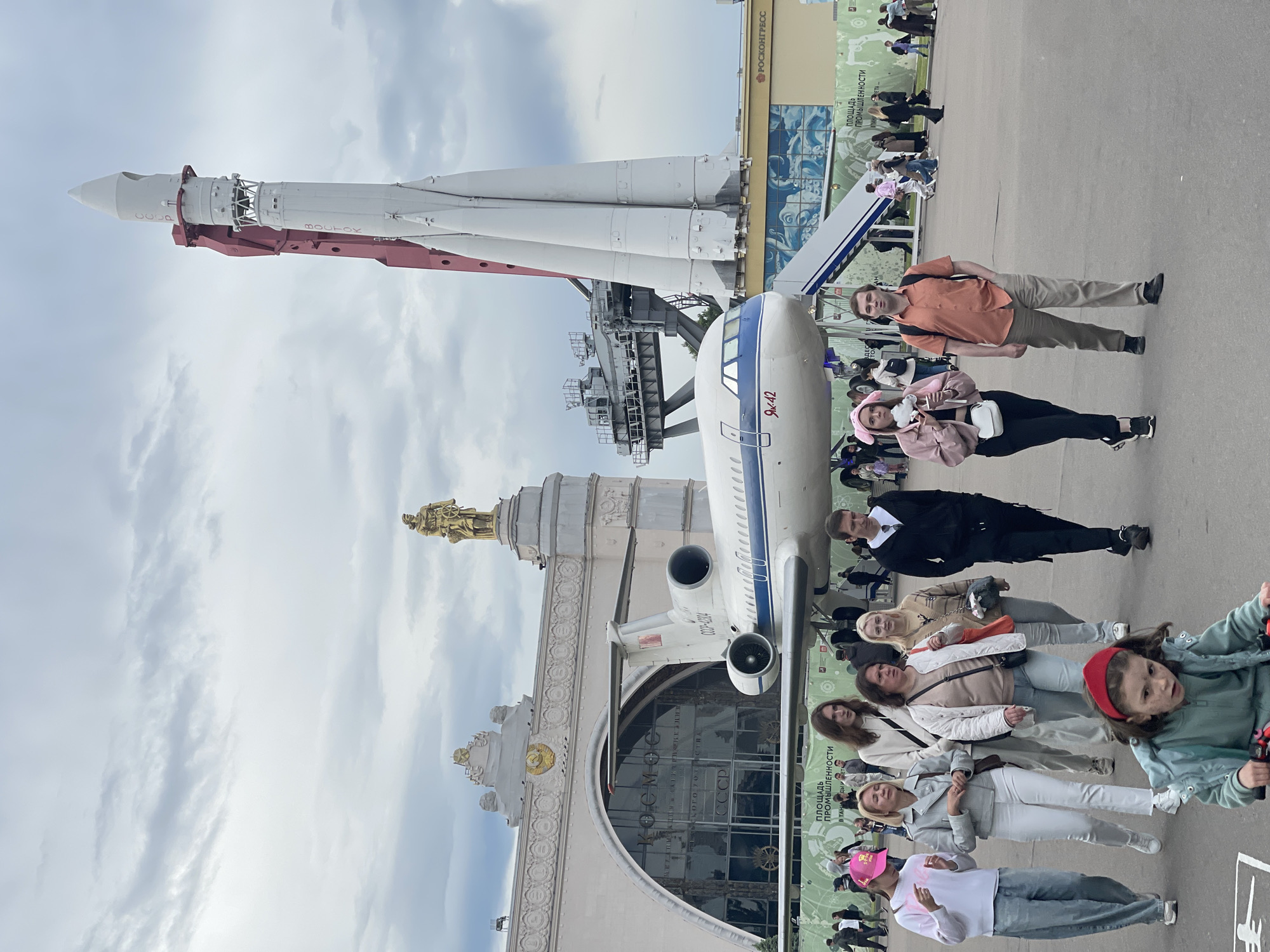 The image shows a group of people standing in front of a large exhibition with a rocket and a plane. The exhibition is located in the city of Unknown, Unknown. The rocket is white with red accents and is mounted on a metal structure. The plane is white with blue stripes. The people are of various ages and are dressed in casual clothing. They appear to be tourists visiting the exhibition. The sky is cloudy.