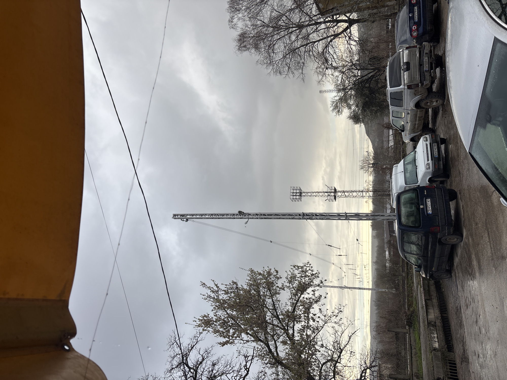 The image depicts an outdoor scene in Cherven Bryag, Bulgaria, under an overcast, grey sky. The perspective is from beneath a yellow corrugated metal awning or overhang, from which water droplets are visible.
The foreground consists of a wet, unpaved or gravel parking area. Parked vehicles include, from left to right: a dark blue Peugeot Partner minivan (license plate CB 4121 AO), a white Renault Kangoo or similar small van (license plate EH 0142 BB), a silver/grey Nissan Navara pickup truck with a bed cap, appearing modified with lifted suspension and off-road tires, splattered with mud (license plate EH 9000 KT), and a dark blue Volkswagen Passat sedan (license plate EH 6178 KC). The front of a fifth silver car is partially visible in the bottom right corner.
Behind the vehicles, railway tracks are visible, accompanied by an overhead electrical catenary system supported by metal poles. Adjacent to the tracks, the ground is covered with bare deciduous trees and brush.
Prominently featured in the mid-ground is a tall, slender metal lattice mast or communication tower, equipped with multiple guy wires and a light fixture. A smaller, similar lattice tower is visible further in the background to the right of the main tower. On the left, a deciduous tree displays sparse, light-colored blossoms or early foliage, while trees on the right are bare.
In the distant background, rolling hills are partially obscured by atmospheric haze or low clouds. No individuals are visible in the scene, and no actions or interactions are depicted.
