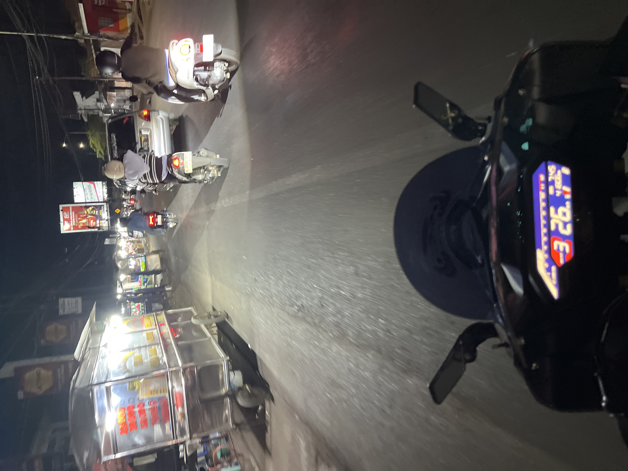 This is a point-of-view shot from a motorcycle at night, traveling down a street in Pecangaan, Indonesia. The road surface is asphalt, and the scene is illuminated by streetlights and the headlights of passing vehicles. To the left of the road, there is a food cart with a stainless steel exterior, displaying prices in red lettering. Further down the street, other food carts and storefronts are visible.
Several motorcycles are present. Directly in front, the rider's motorcycle dashboard displays "26." followed by "7.745" and "4806". To the right, a white scooter is occupied by a person wearing a dark jacket and helmet. In the middle distance, a silver car is visible, and to its left, another motorcycle with a rider in a striped shirt and helmet is in motion. Another motorcycle is seen further ahead, with its rear lights illuminated. Overhead, power lines crisscross the sky, and billboards are mounted on poles. The overall impression is a bustling nighttime street scene with active commerce and traffic.