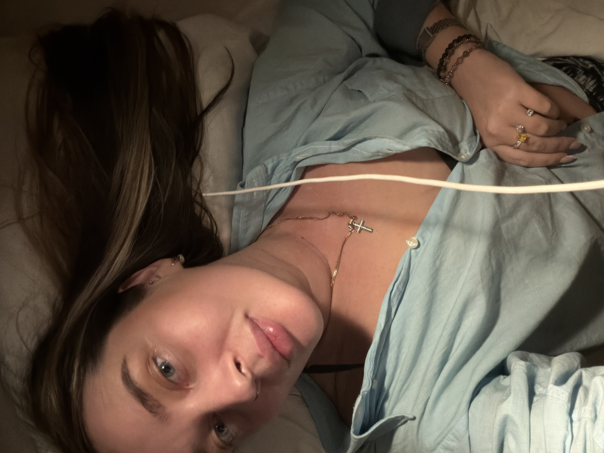 A woman with fair skin and blue eyes is lying down, angled towards the camera. She has long brown hair with blonde highlights. She is wearing a light blue button-down shirt and a gold necklace with a cross pendant. On her right hand, she wears several bracelets and rings. A white wire or cord is visible across her chest. She is resting on a white surface, possibly a pillow or bed. The scene provides no information to determine the location in Moscow, Russia.