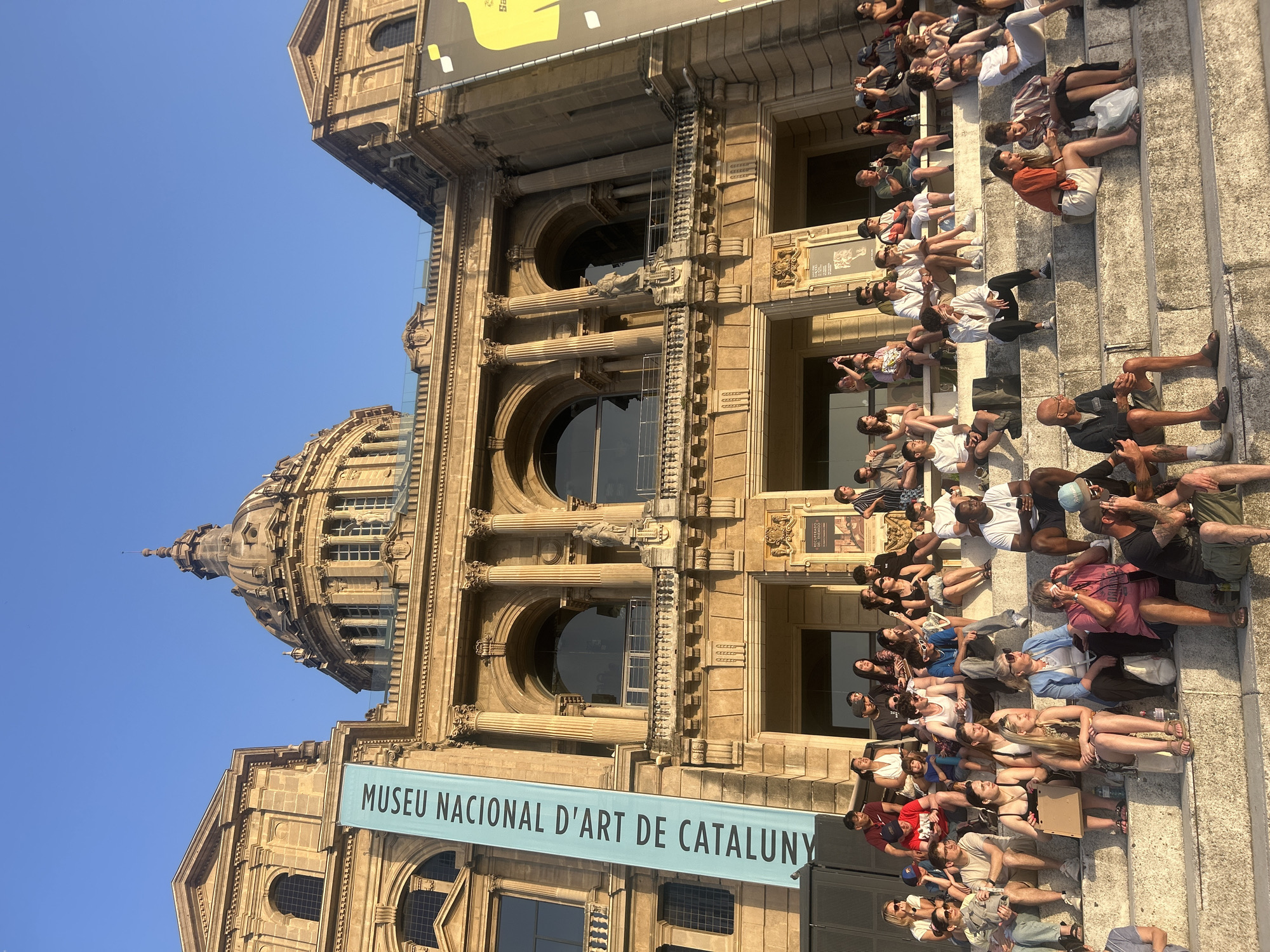 The image depicts a crowd of people sitting on stone steps in front of the Palau Nacional, a grand building in Barcelona, Spain, which houses the Museu Nacional d'Art de Catalunya. The building features classical architecture with columns, arches, and a prominent dome. A large blue banner with white lettering, reading "MUSEU NACIONAL D'ART DE CATALUNYA," hangs vertically on the facade. The sky is clear and blue, suggesting it is daytime. The people are diverse in age and are casually dressed, many looking towards the building or engaging with each other. Some individuals are holding phones, possibly taking pictures or videos. The overall scene conveys a sense of public gathering and appreciation for the historic architecture and museum.
