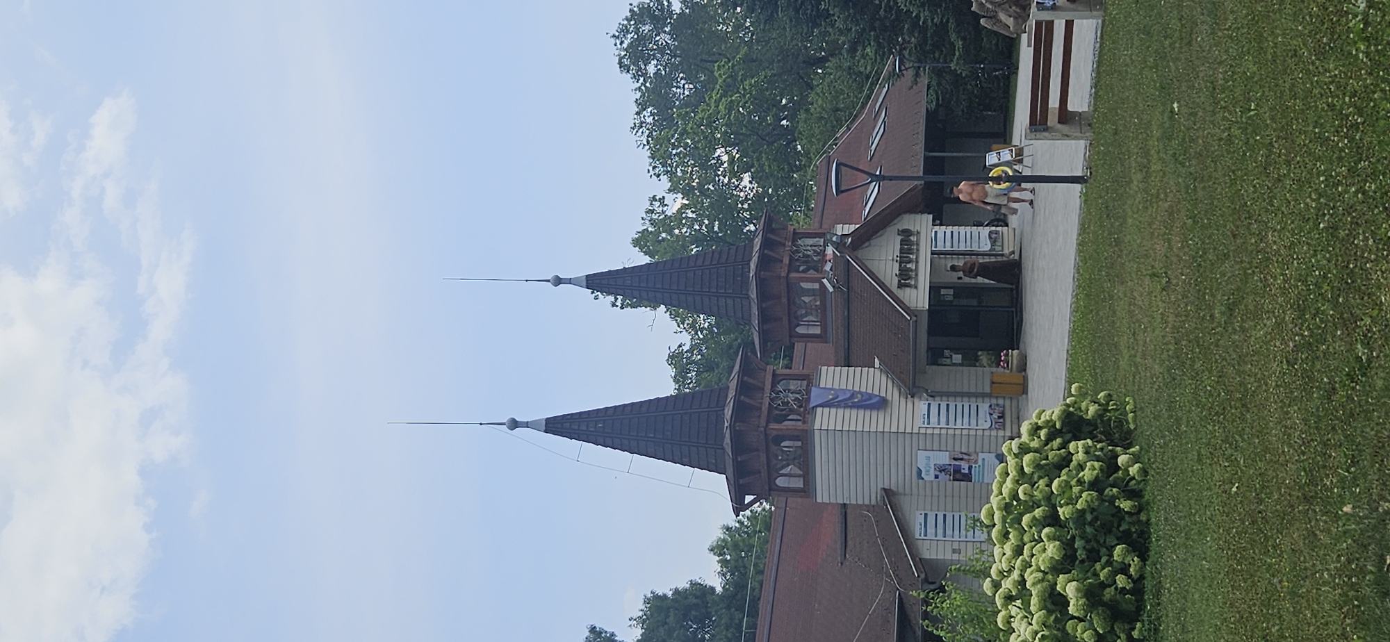 The image shows the entrance to the Tófürdő bathhouse in Hévíz, Hungary. The building features a distinctive architectural style with two prominent conical towers topped with spires. The facade is a mix of light-colored siding and dark wood accents, with a reddish-brown tiled roof. A sign above the entrance reads "TÖFÜRDŐ".
In front of the building, there is a paved area with several people. One person, a male wearing shorts, is walking away from the entrance, carrying a bag. Another person, a female in a long dress, stands near the entrance. A large, bushy green plant is in the foreground, to the left of the entrance, with a grassy lawn and a park bench visible to the right. The scene is set against a backdrop of lush green trees and a pale blue sky with scattered clouds.