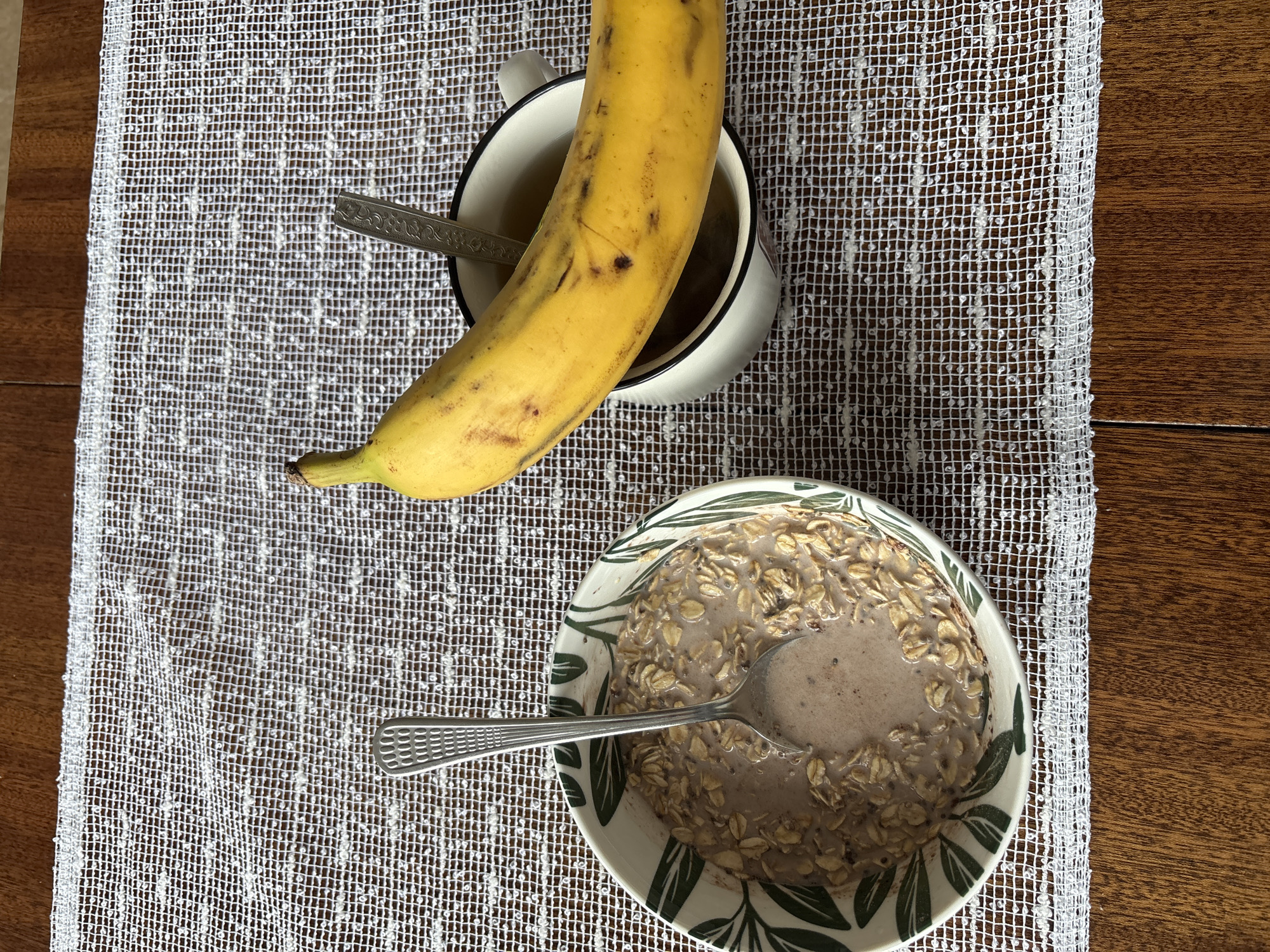 A high-angle shot captures a breakfast scene set on a dark wooden table partially covered with a white, textured placemat. In the lower part of the frame, there is a bowl of oatmeal with a spoon resting inside. The bowl has a white base adorned with green leaf-like designs around the rim. Above the bowl, there is a white mug containing a dark liquid and a small spoon. A yellow banana rests atop the mug. The placemat beneath the items displays a woven, net-like pattern, contrasting with the smooth surface of the wooden table. No people are visible. The location is not discernible from the image.