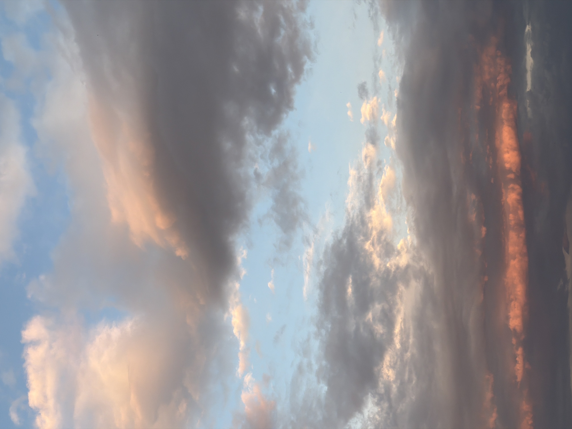 The image shows a view of the sky with various cloud formations. The sky is visible in the gaps between the clouds, appearing as a light blue color. The clouds range in color from light grey to dark grey, with some clouds showing orange and pink hues, likely reflecting the light of the setting sun. The image is taken from Petrovac na Moru, Montenegro.