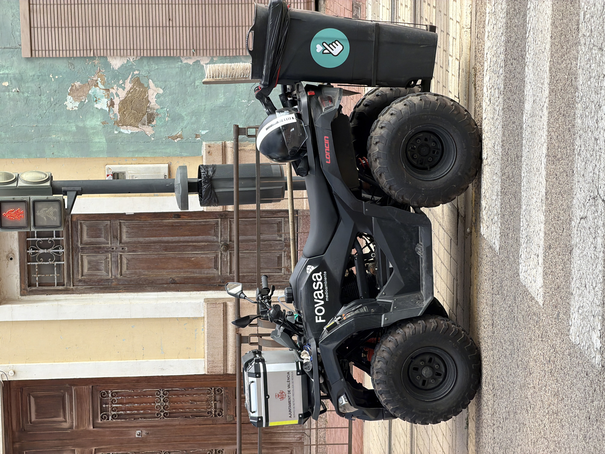 An all-terrain vehicle (ATV) is parked on a city street, facing left. The ATV is black and equipped with a storage box on the front rack, a helmet resting on the seat, and a large black trash bin attached to the rear rack. The storage box on the front has a logo that reads "AJUNTAMENT DE VALENCIA SERVICIO DE NETEJA Y RECOLLIDA DE RESIDUS". A sticker with a hand forming a heart shape within a teal circle is affixed to the trash bin.
The background features a building with two large wooden doors, a yellow facade, and a distressed green wall. A traffic light and a trash can are also visible in the background, to the left of the ATV. The street appears to be paved with cobblestones and has a pedestrian crossing marked by white stripes. The overall environment suggests an urban setting, possibly in Spain due to the text on the storage box.