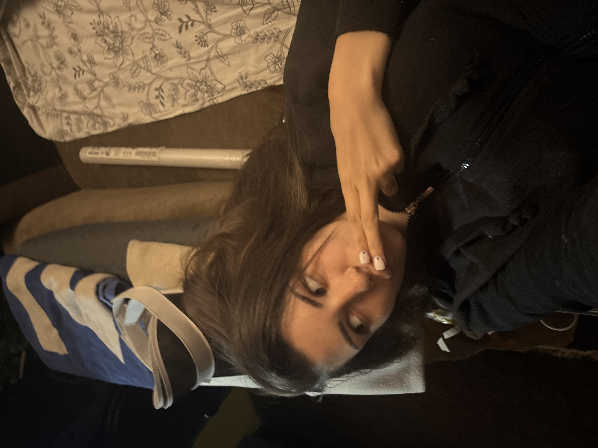 The image portrays an indoor scene, likely a bedroom or living room, characterized by dim lighting, suggesting evening or nighttime. The central subject is a young woman with dark brown hair, seen reclining or lying down on a piece of furniture, possibly a couch or bed. She is wearing a black long-sleeved top or hoodie, which features a visible zipper and drawstring details. Her right hand is positioned near her mouth or chin, with fingers slightly curled, and her fingernails are painted a light color, possibly white or pale pink, each adorned with a small, darker dot. She looks towards the right side of the frame with a neutral to subtly pensive expression.
In the upper part of the image, behind her head, is a light-colored sheet or duvet cover patterned with a grey or black floral and botanical design. Below this, a long, white cylindrical object, possibly a tube for a poster or artwork, is visible. It bears small, indistinct text and symbols on its surface, which are not clearly legible. Further to the left and foreground, a blue and white striped bag or fabric item with prominent white handles rests on dark-colored furniture. The overall composition suggests a relaxed, personal moment within a residential setting in Belopoptsi, Bulgaria, though no specific geographical identifiers are present within the visual content. The original image appears rotated 90 degrees counter-clockwise.