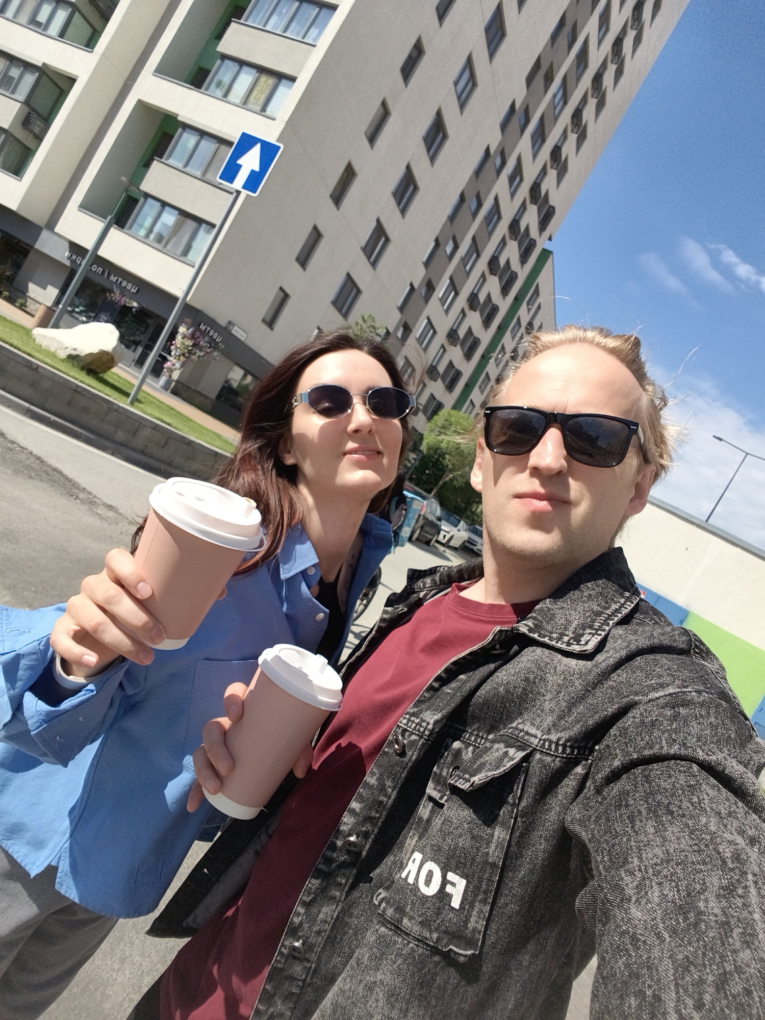 This bright outdoor selfie features two individuals, a woman and a man, on a sunny day in what appears to be an urban setting in Tyumen, Russia. Both are holding disposable coffee cups and posing for the camera.
The woman is on the left, with long dark brown hair and wearing silver-framed sunglasses. She has a soft smile and is dressed in a light blue collared shirt and grey pants. Her right hand extends forward, holding a light pink or beige coffee cup with a white lid.
To her right, the man has blonde hair and wears rectangular black sunglasses. He holds an identical coffee cup in his left hand, also extended towards the camera. He is wearing a dark red t-shirt under a dark grey, acid-wash denim jacket and has a neutral expression.
The background features tall, modern-looking buildings with numerous windows and balconies, displaying a contemporary architectural style. The buildings are predominantly light-colored, with some green accents visible in the balconies. A paved road or sidewalk is in the mid-ground, alongside a grassy area with a large white rock sculpture and some flowering plants. A blue road sign with a white upward arrow is prominently visible near the buildings. The sky is clear and blue with minimal clouds, indicating bright, sunny weather and daytime.
Visible text includes "ЯОF" (which reads as "FOR") in white letters on the pocket of the man's denim jacket. On a shopfront window in the background, reversed and somewhat blurry Cyrillic text, such as "нкя on | мтбэи", can be faintly discerned.