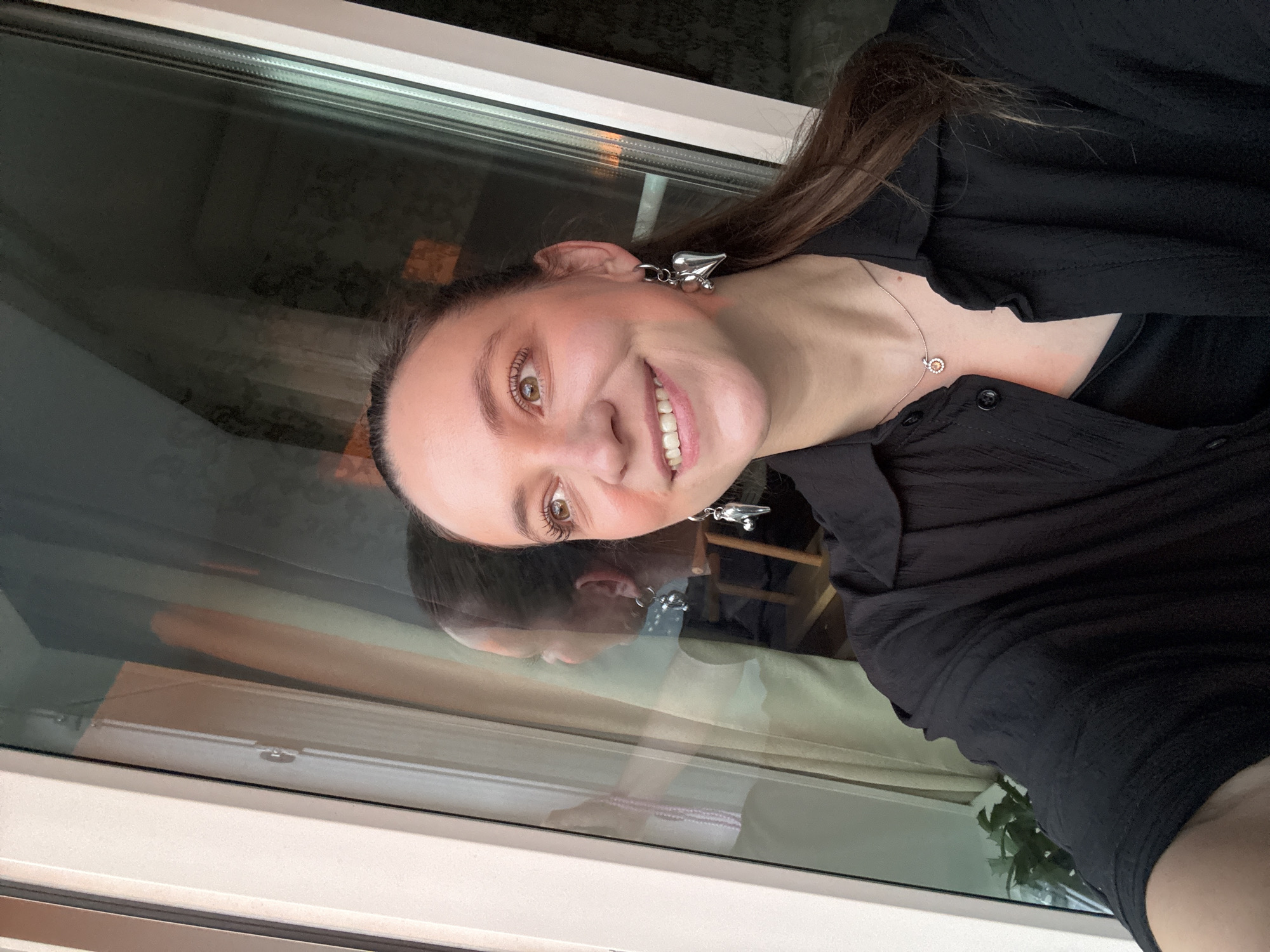 A smiling woman wearing a black button-down shirt and silver earrings is seen in a selfie. She has brown hair and brown eyes. She is wearing a necklace. Reflected in the window behind her is a similar image, indicating a reflection in the glass. The surrounding environment and city of Chaikovsky, Russia cannot be determined from the available visual information.