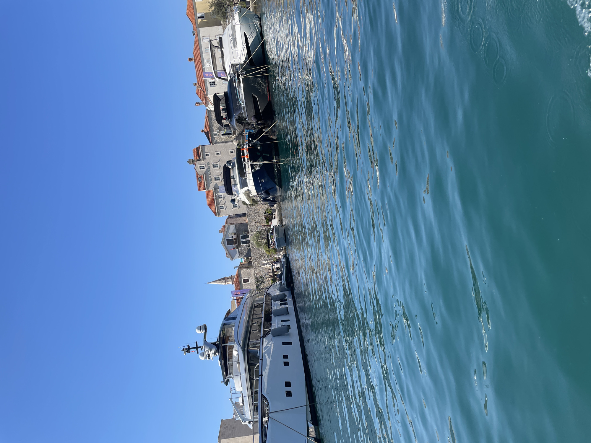 A wide-angle, eye-level shot captures a serene scene in Budva, Montenegro, with the azure Adriatic Sea dominating the foreground and reflecting the clear blue sky. In the background, a line of moored yachts rests peacefully against the backdrop of Budva's historic Old Town. The prominent white yacht on the left boasts a sleek, modern design, while three other vessels—a dark blue, a black, and a white yacht—are clustered together to its right. Behind the yachts, traditional red-tiled roofs and stone buildings of the Old Town are visible, with a slender church spire piercing the skyline. The water's surface is gently rippled, with subtle circular patterns hinting at the movement of unseen forces. The overall atmosphere is one of tranquility and vacation allure.