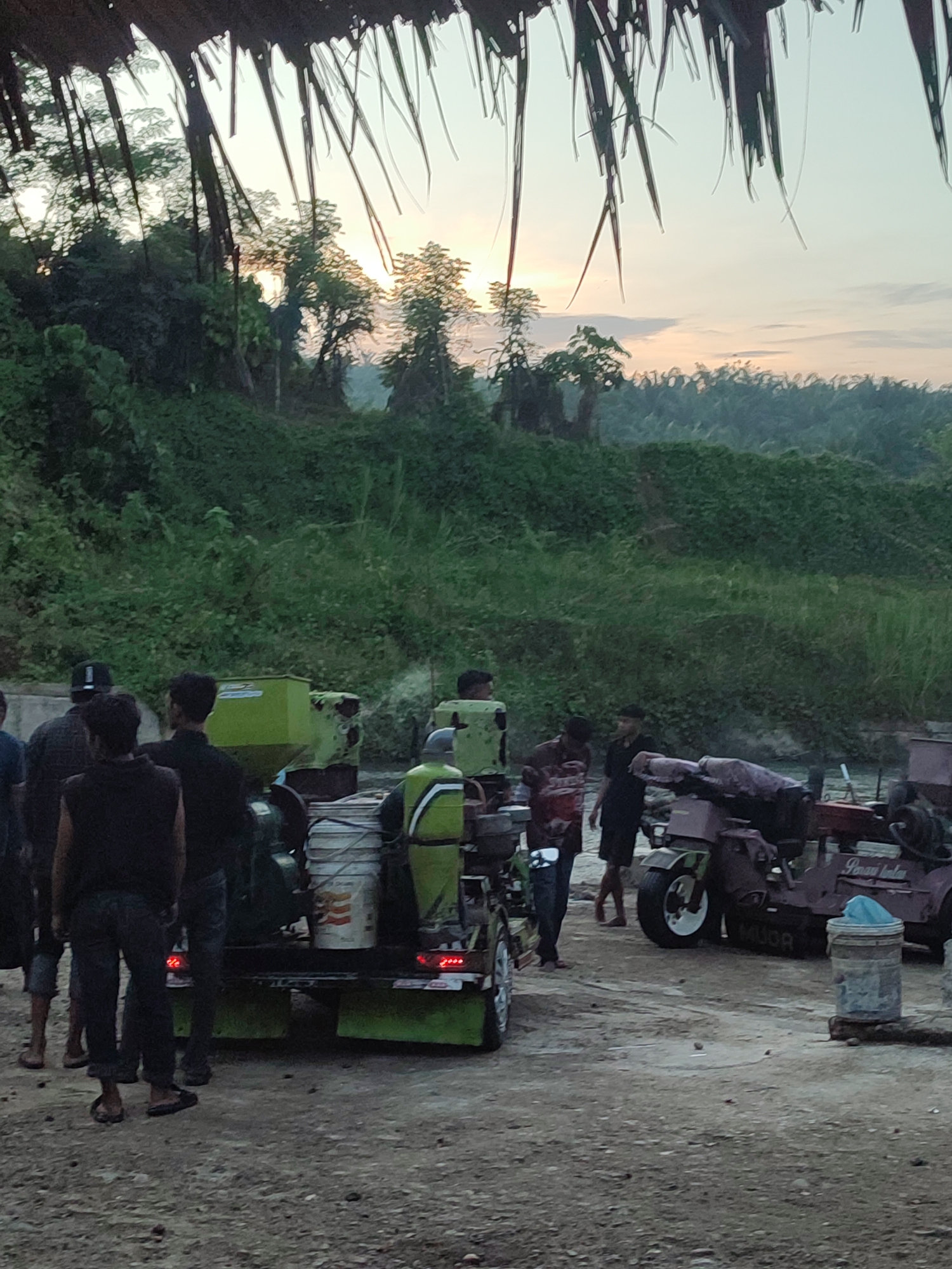 The image depicts an outdoor scene at what appears to be dawn or dusk, with a soft, diffused light illuminating the sky and landscape. In the foreground, a rough, gravelly surface is visible, with several individuals gathered around a couple of mechanical devices. The overall impression is one of an industrial or agricultural setting, possibly a rural area on the outskirts of Saweuk, Indonesia.
In the center-left of the image, a bright green contraption, likely a processing machine or generator, is mounted on a trailer. Several people are standing nearby, some appearing to be working or observing the equipment. One individual in the foreground wears a dark sleeveless shirt and jeans, facing away from the camera. To their right, another person in dark clothing stands. Further back, a person in a green and yellow vest is seen near another piece of machinery.
To the right, a reddish-brown motorized vehicle, resembling a modified scooter or small tractor, is parked. It has a large rear wheel and a seat covered with fabric. Text is visible on the side of this vehicle, appearing to read "MUOR" and "Pinag kates." A blue plastic bag is placed on a cylindrical object next to this vehicle.
The background features a dense, verdant hillside covered in lush vegetation, with trees interspersed throughout. The sky above is a pale gradient of yellow and white, suggesting the presence of the sun just above the horizon. Hanging thatch or palm fronds are visible at the very top of the frame, suggesting a covered area or shelter. The overall atmosphere is calm and quiet, with a sense of work being done in a natural environment.