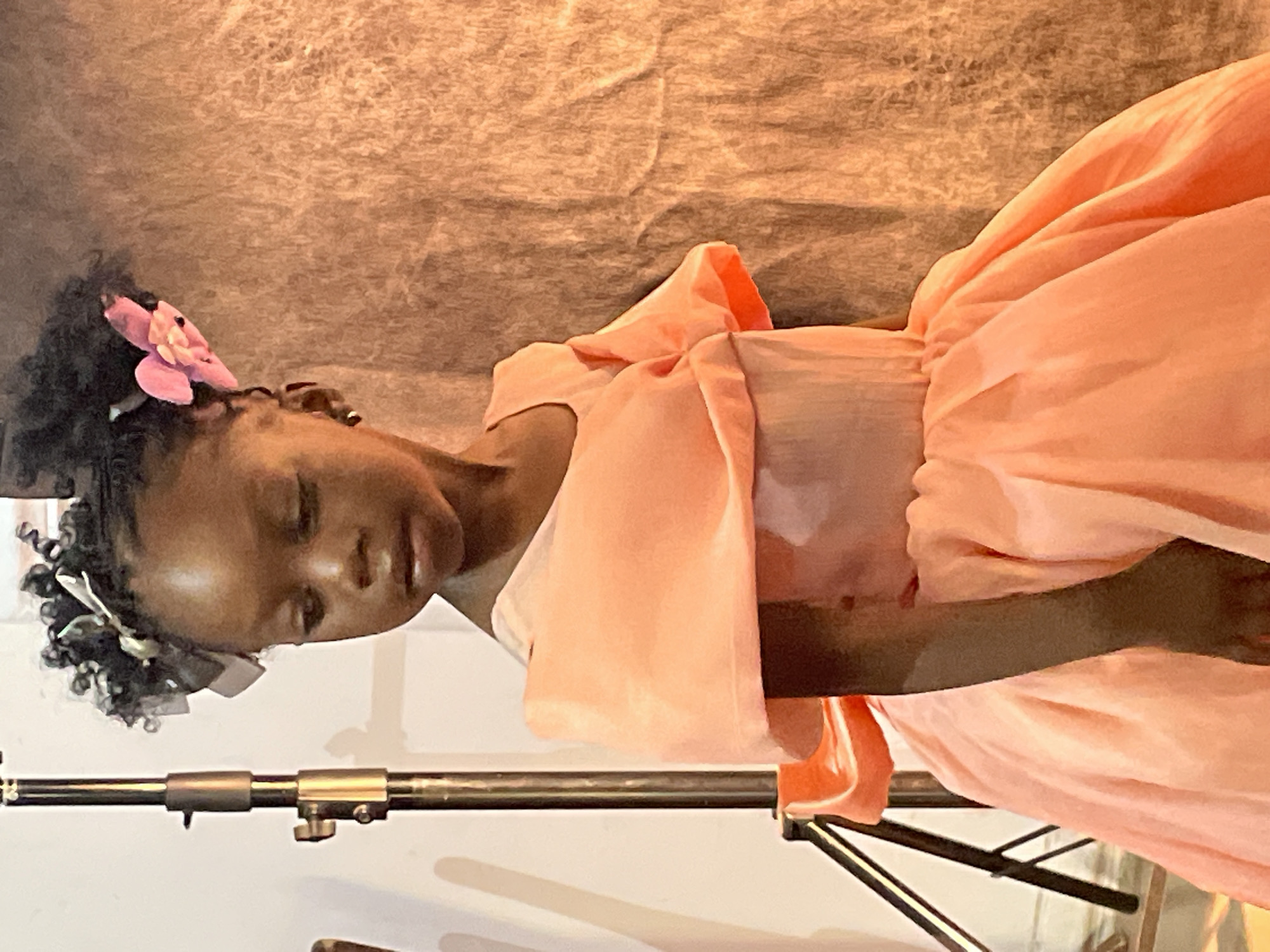 The image, rotated 90 degrees counter-clockwise, captures a young girl in what appears to be an indoor portrait session. The main subject is a young girl, likely of African descent, with dark, curly hair adorned with several small white or silver accessories and a prominent pink fabric flower on the left side of her head.
She is dressed in an elegant, light peach or coral-colored gown featuring puffed sleeves and a voluminous skirt with visible folds and gathers. Her eyes are closed or cast downwards, conveying a serene, calm, and contemplative expression.
The girl is positioned against a light brown or beige textured fabric backdrop, which displays wrinkles and folds, suggesting a professional studio environment. In the lower left quadrant of the image, a metallic tripod stand is partially visible, indicating photographic equipment. There is no visible text in the image. This scene takes place in Jos, Nigeria.