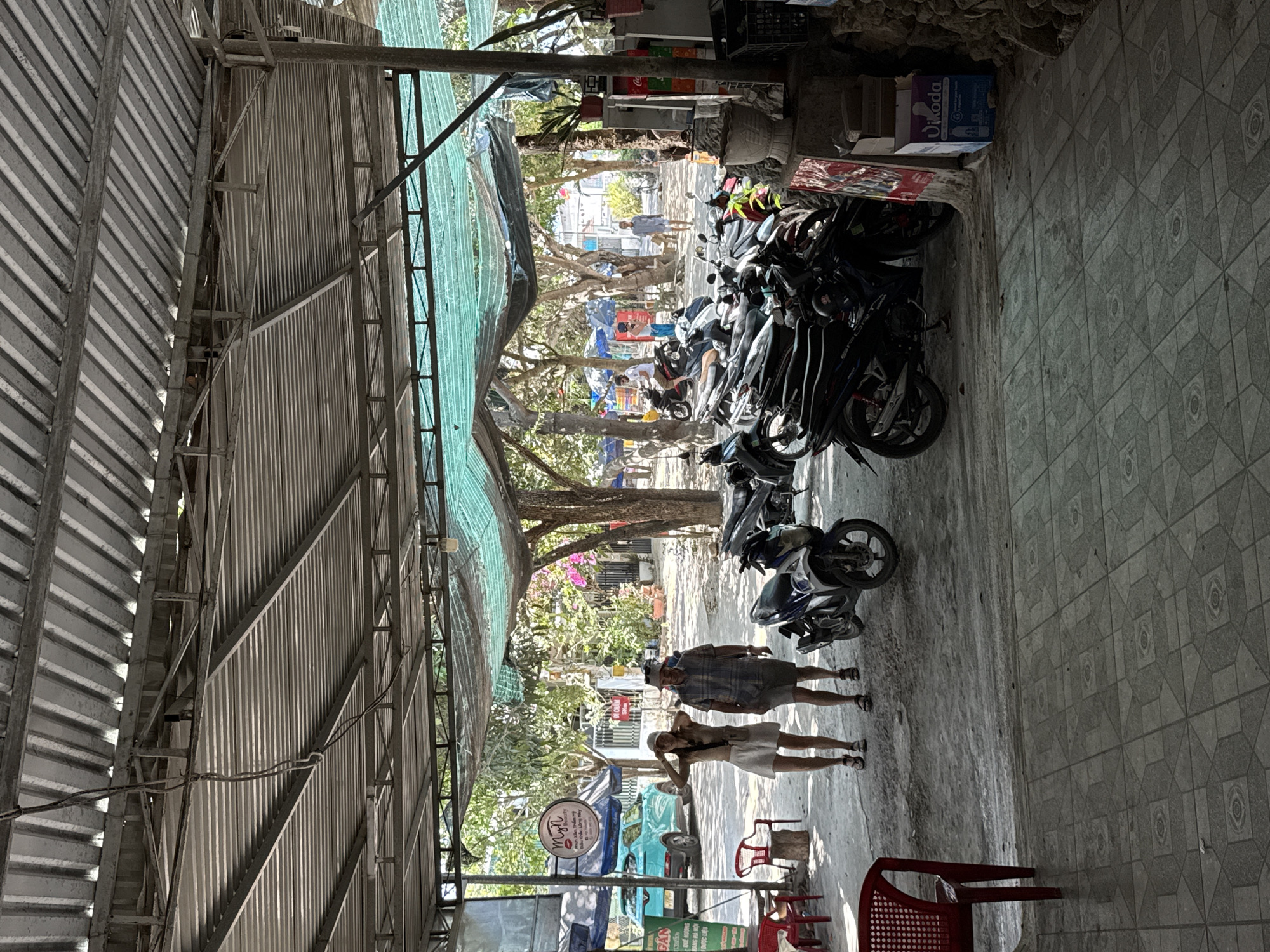 The image shows a street scene in Diên Khánh, Vietnam. The view is taken from under a corrugated metal roof structure, partially covered with a green tarp. Numerous motorcycles are parked haphazardly on the right side of the frame. Two people, a man and a woman, are walking along the street. In the distance, trees line the street, and various commercial signs are visible. Red plastic chairs are present near the bottom of the frame, suggesting a small outdoor dining area. The overall scene conveys a typical streetscape in a Southeast Asian town.