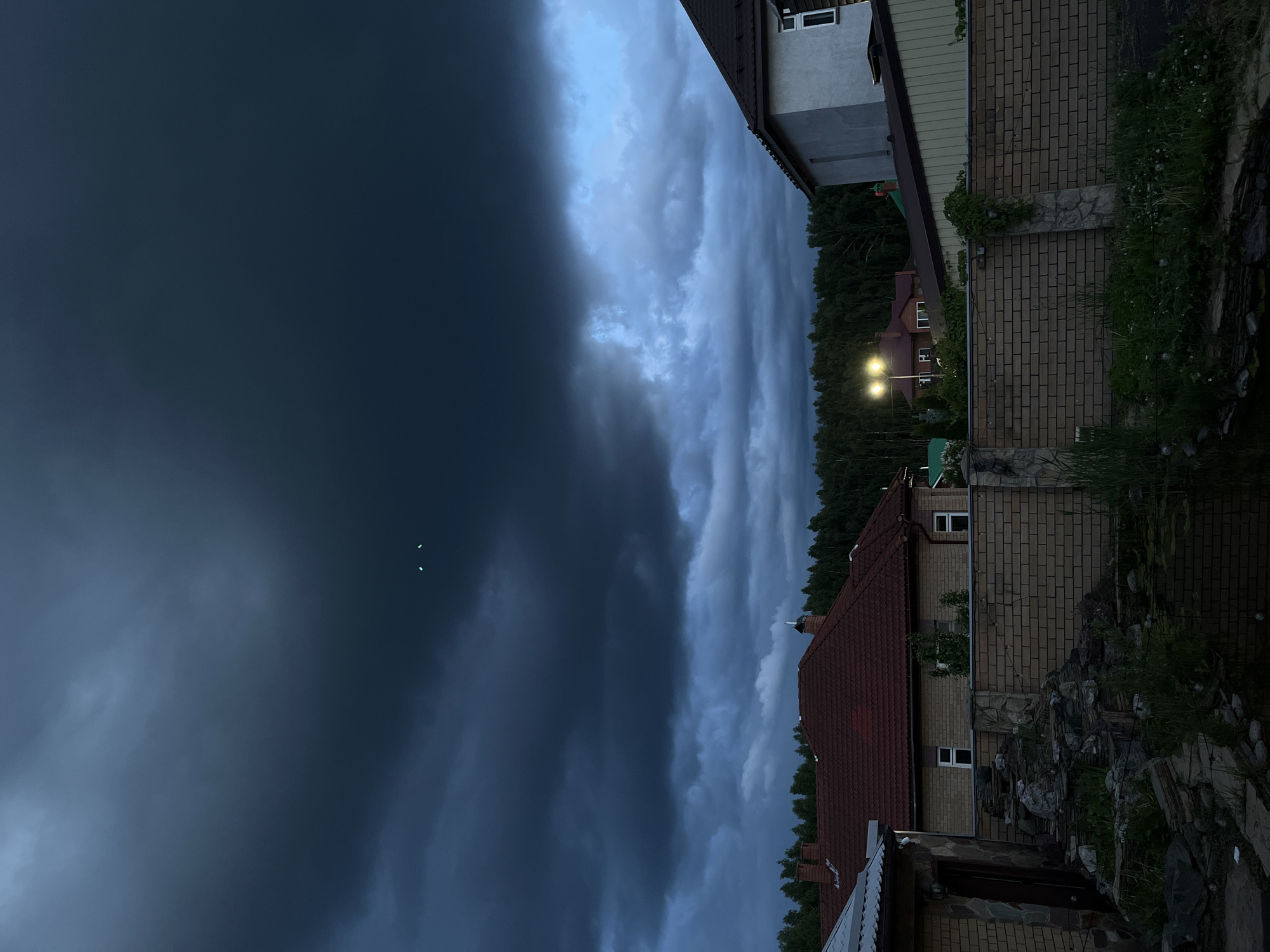 The image captures a residential scene in Ореховка, Russia, at what appears to be dusk or night due to the low light and illuminated structures. The sky is a prominent feature, covered by heavy, dark clouds. The left portion of the sky is a deep, uniform blue-black, while the right features turbulent, lighter grey-blue clouds, suggesting dramatic or stormy weather. Two small, bright white specks are visible in the darker part of the sky, possibly birds or distant lights.
In the mid-ground and foreground, several houses are visible. On the right, a house with a brick base and a lighter-colored upper story with windows is partially seen, topped by a dark-tiled roof. Further into the frame, another house with a distinctive red-tiled roof and brick walls is visible, illuminated by bright outdoor lights. A dense line of dark green forest acts as a natural backdrop behind the houses. The immediate foreground includes grassy areas, some small plants, and a rocky landscaping feature near the houses. No people or visible text are present. The scene conveys a quiet, possibly impending weather atmosphere.